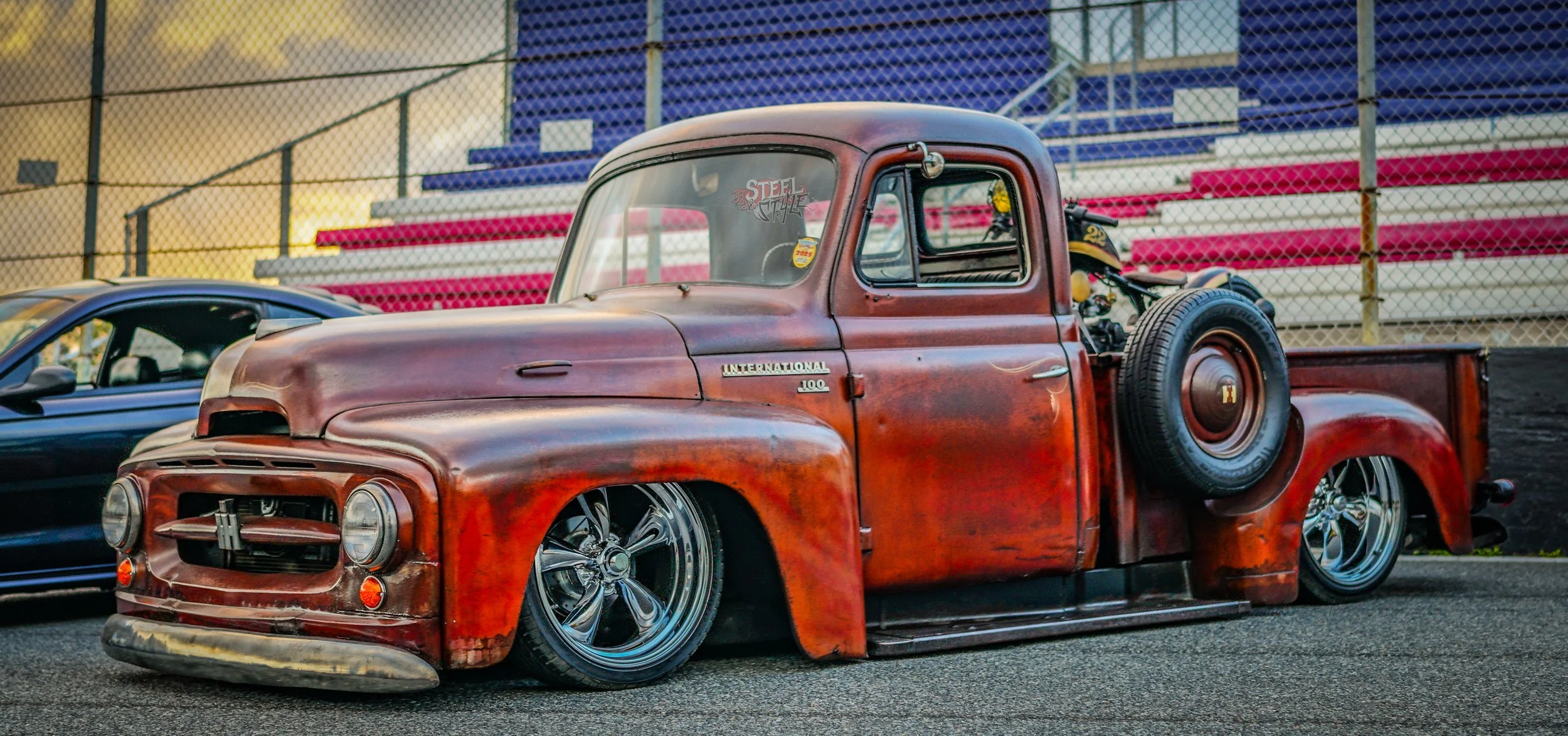 A vintage red pickup truck with a weathered rust finish and lowered stance parked on a race track with a motorcycle in the truck bed and a blue sports car nearby, with stadium seating and a chain-link fence in the background.