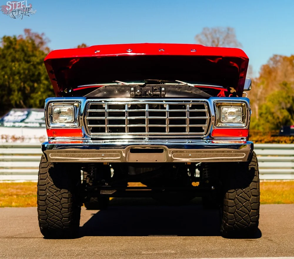 Front view of a vintage red Ford pickup truck with its hood open, showing the grille and headlights, parked on a paved surface.