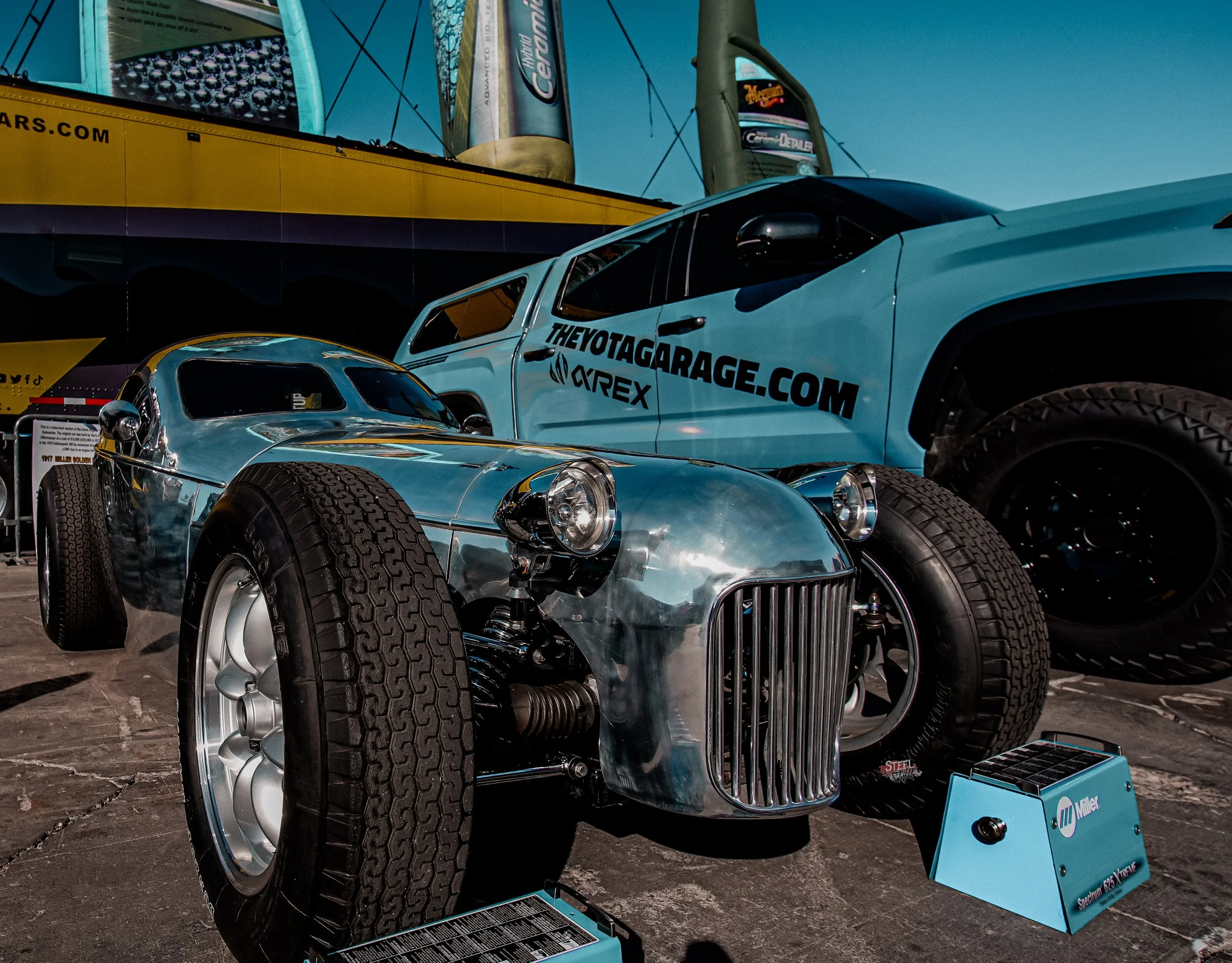 Vintage race car with a reflective metal body and large tires displayed at an outdoor auto event, with a large sponsor truck and billboard in the background.