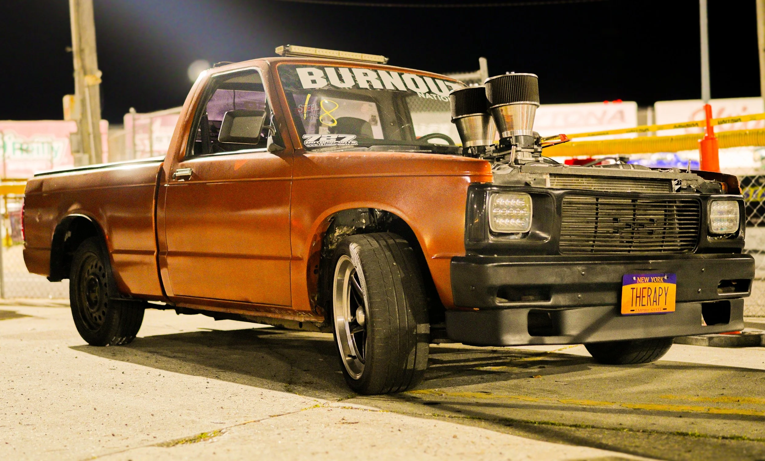 A vintage brown pickup truck with custom modifications, including a large engine protruding from the hood and a live of fire on the front license plate that reads "THERAPY," parked at night in a lot with a chain-link fence and colorful signs in the b
