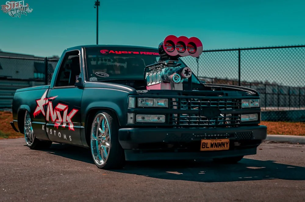 Customized black pickup truck with large chrome wheels, a decal on the side that reads 'FUSK KUSTOMS', the front grille removed, a large supercharger with three red circular air intakes protruding from the hood, and a custom license plate that says '