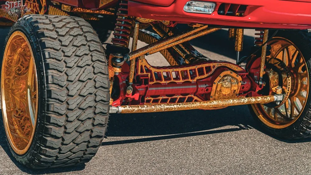 Close-up of a customized vintage car's front wheel and suspension, with gold-colored wheels, red and gold chassis, and thick treads on the tire.
