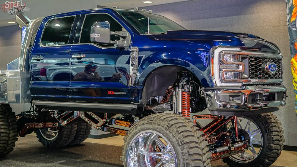 A large, modified Ford truck with a blue body, oversized off-road tires, and lifted suspension on display indoors. The truck has custom components, including a metal frame and orange coil springs visible on the front suspension.
