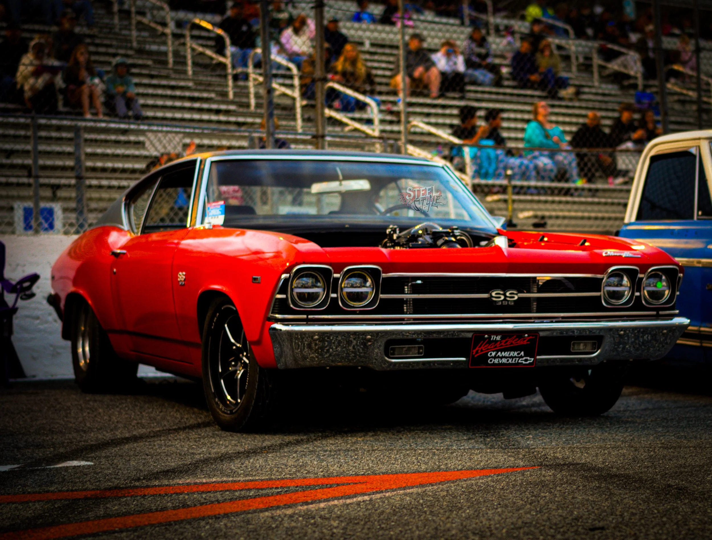 Red vintage Chevrolet SS 396 muscle car parked in a racing pit area with spectators in the stands behind a chain-link fence.