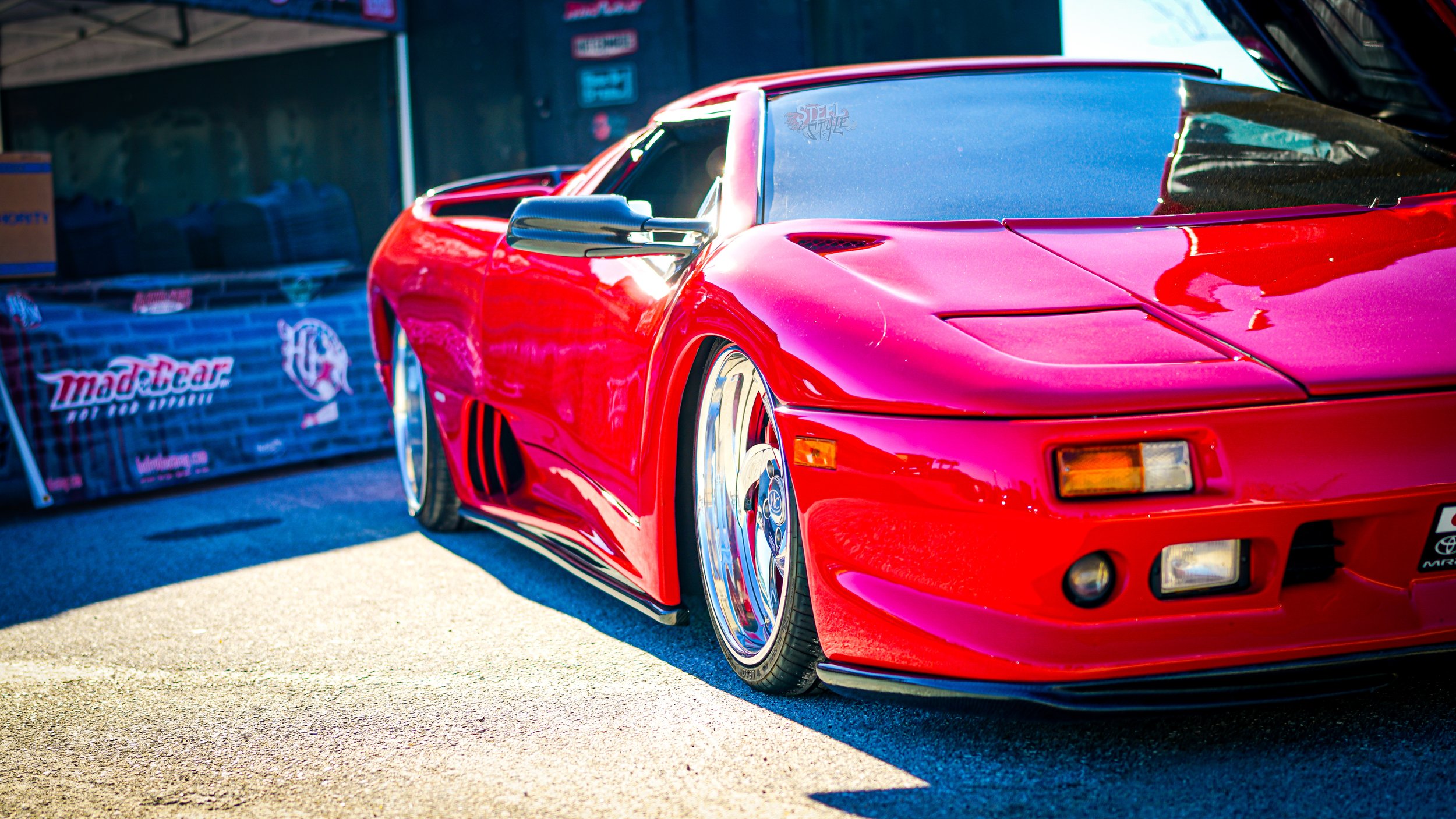 Red sports car with aftermarket wheels parked outdoors near a black barrier with MadeCar branding.