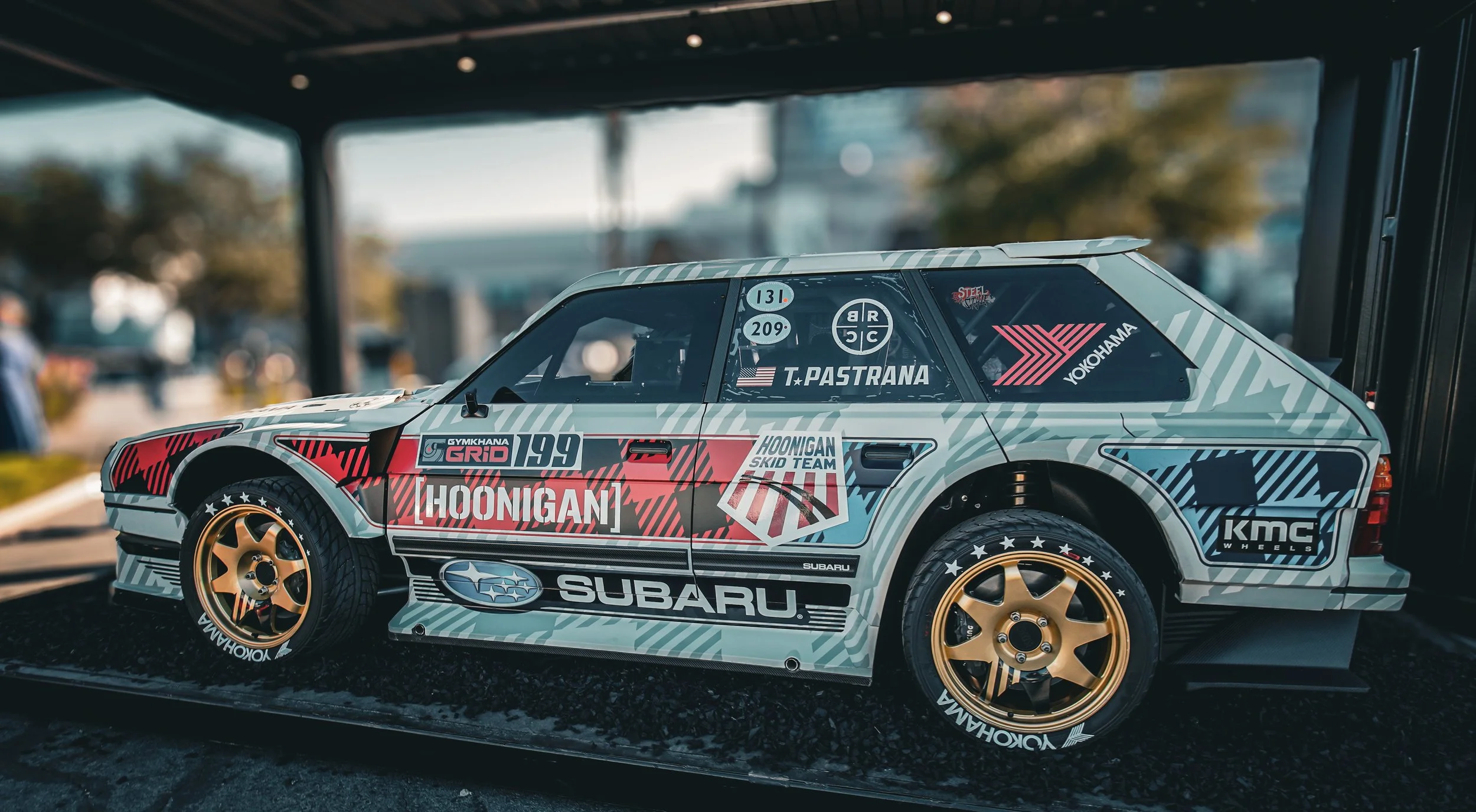 A detailed scale model of a vintage Subaru race car with colorful sponsor decals, gold wheels, and racing tires, displayed on a black platform.