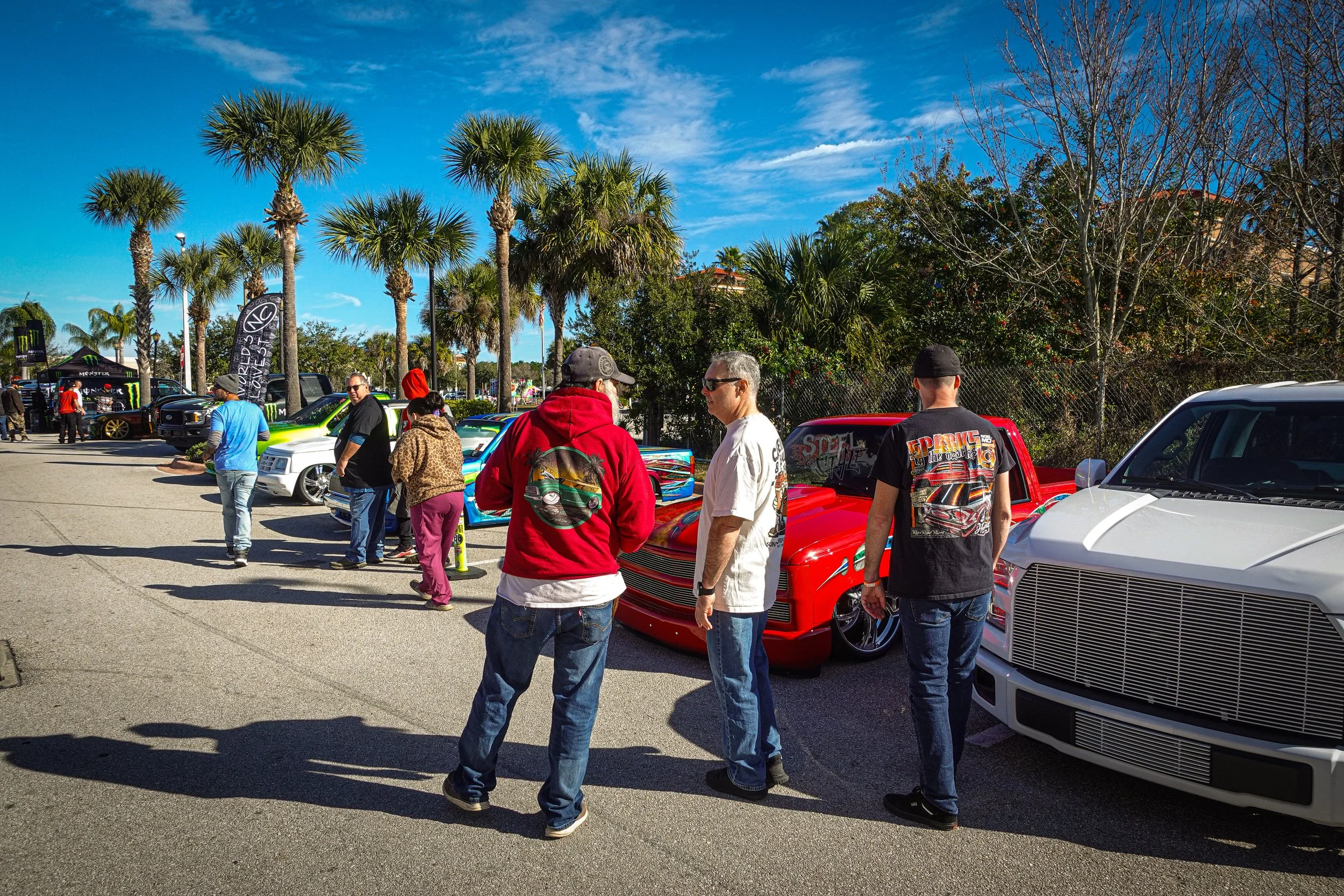 Line of customized cars at a car show on a sunny day with palm trees and blue sky in the background.