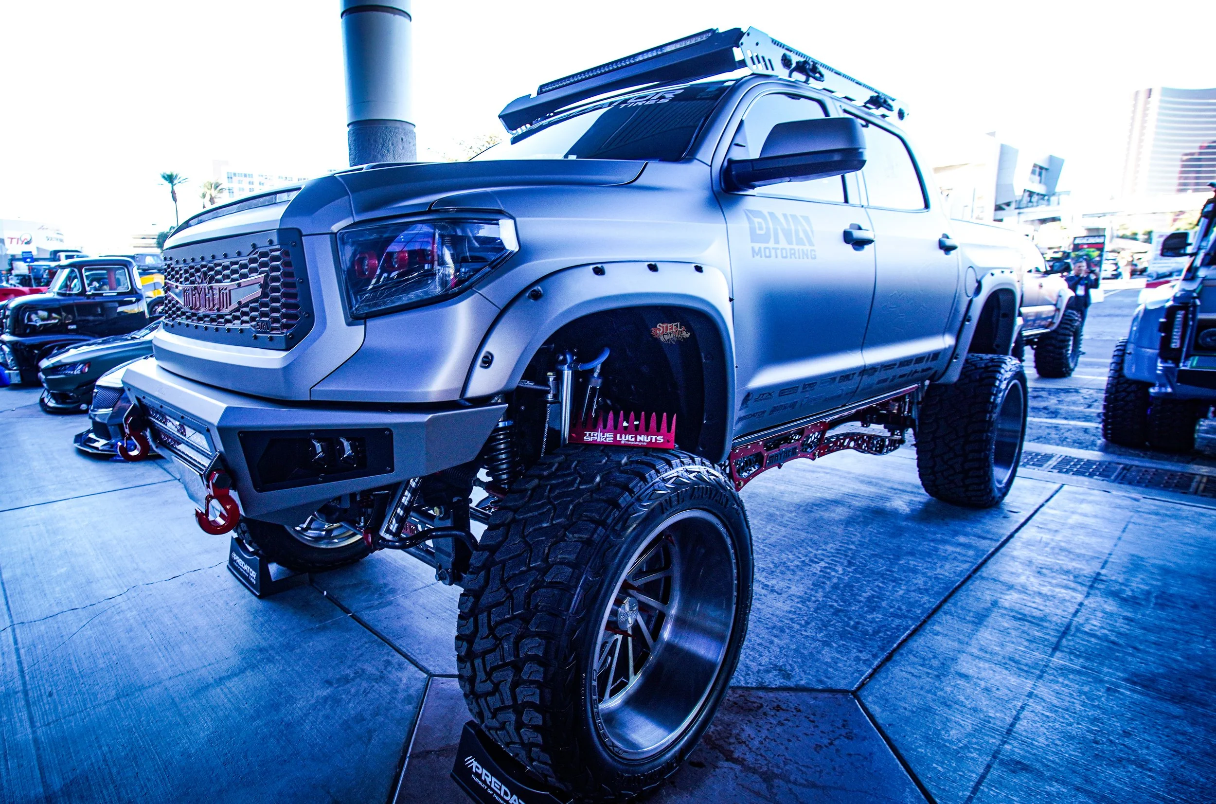 Modified pickup truck with large off-road tires and suspension, aftermarket grille, and custom equipment mounted on the roof, displayed at an outdoor car show.