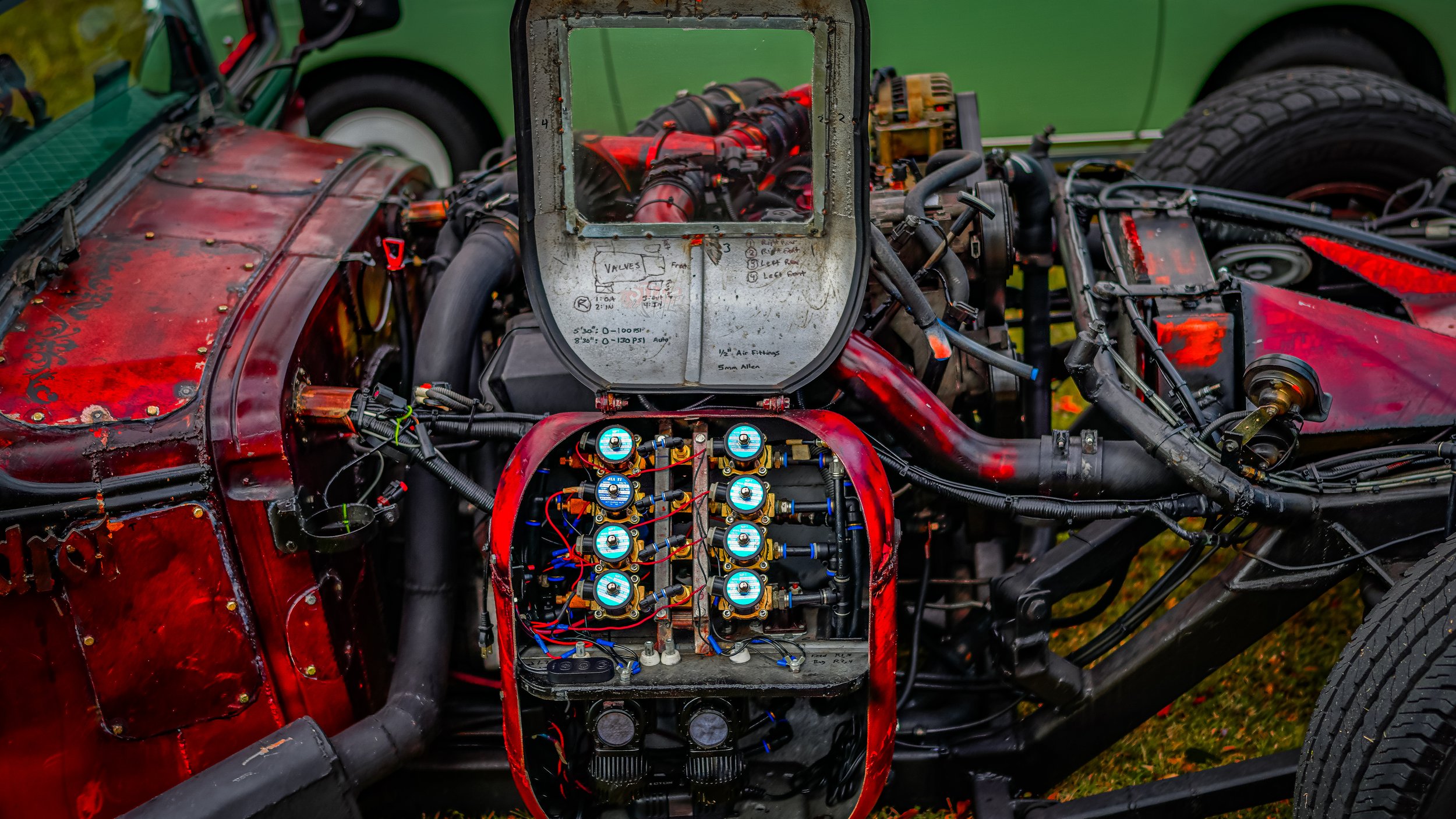 Close-up of a vintage race car engine with a red, weathered body and exposed mechanical components, including wiring, hoses, and gauges.