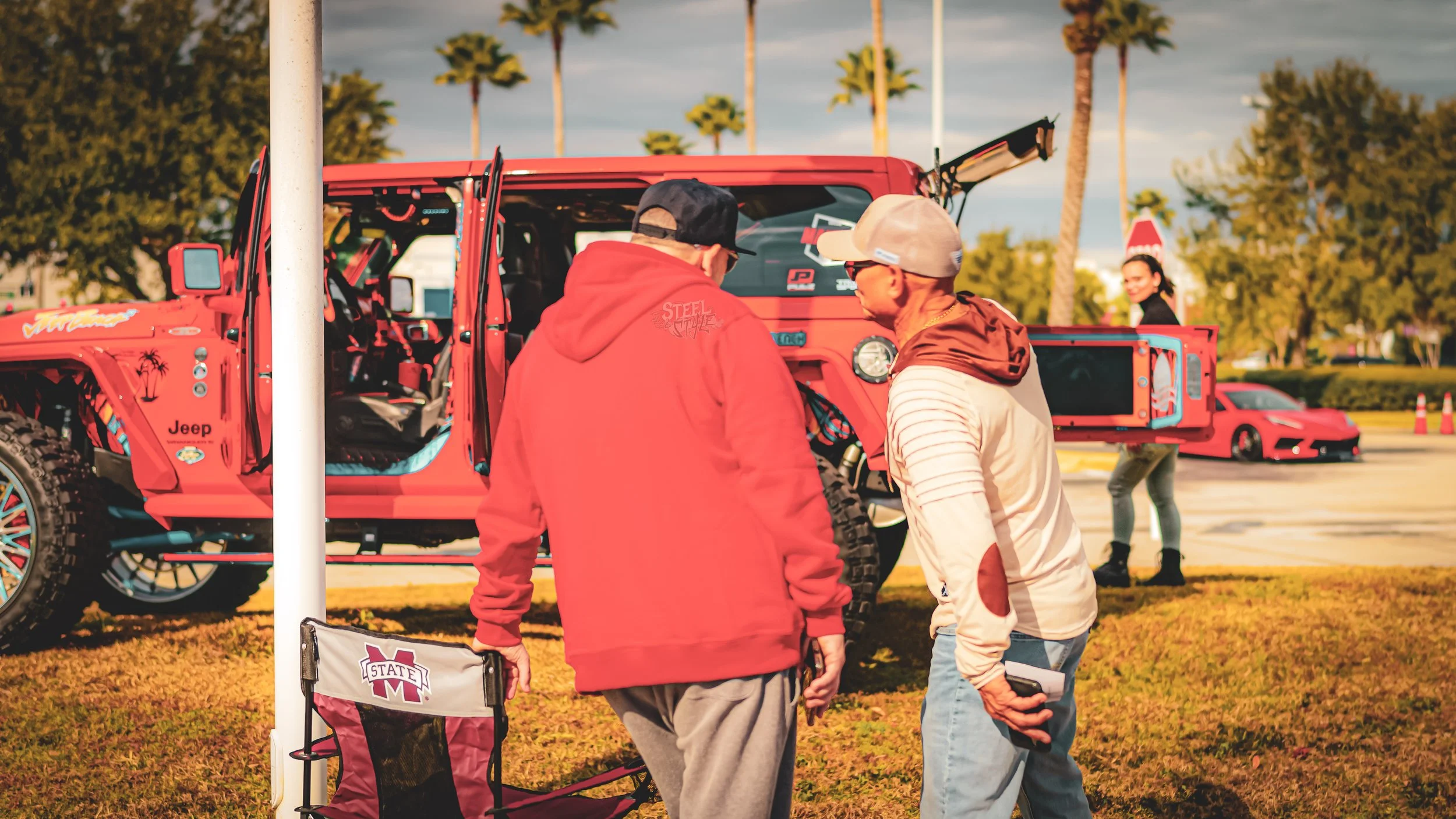 Two men are standing in front of a red off-road Jeep, engaged in conversation, at an outdoor event with palm trees, a red sports car, and a smiling woman in the background.