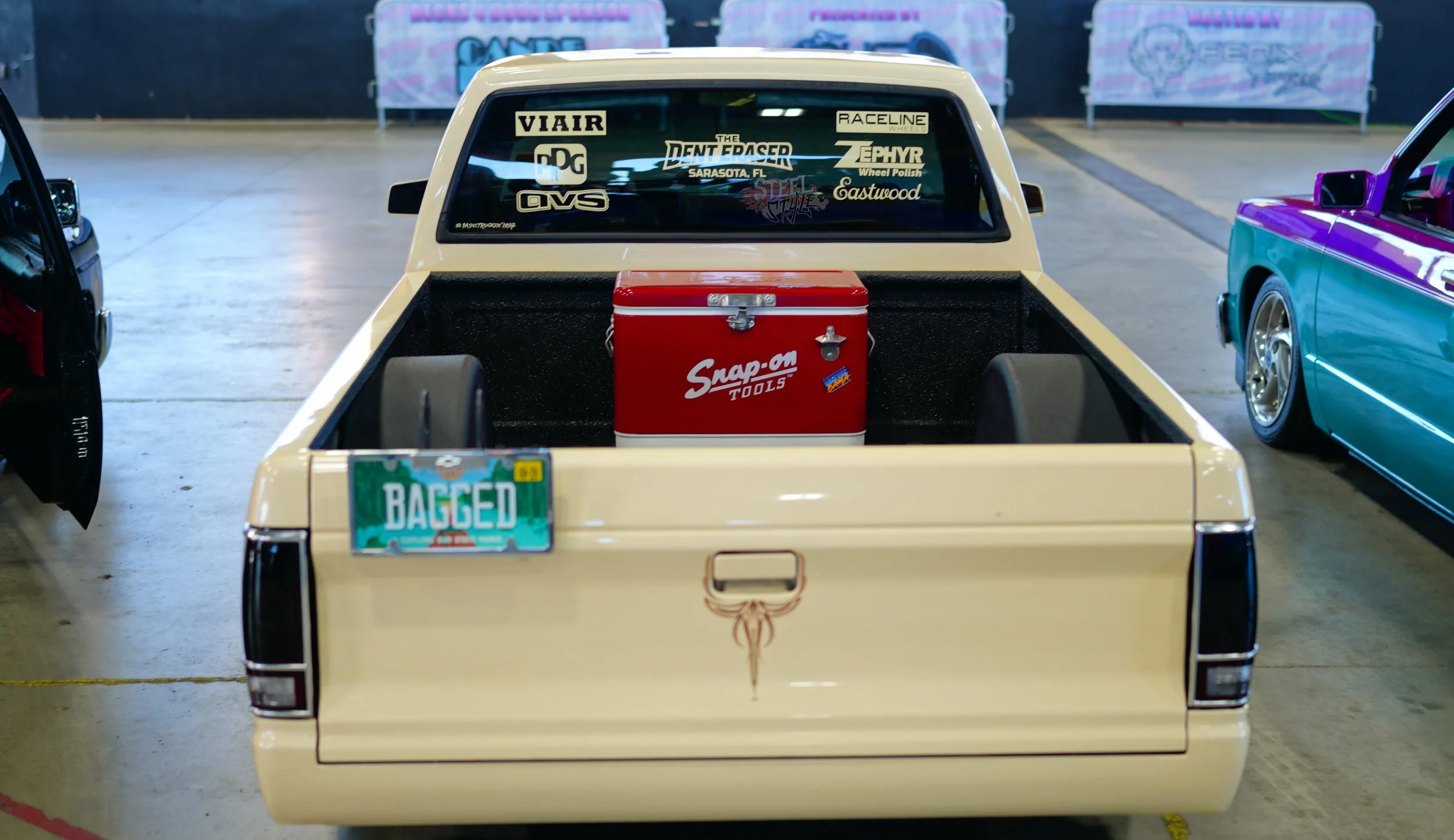 Rear view of a vintage pickup truck with various stickers on the back window and a red toolbox in the truck bed, inside an indoor car show or exhibition.