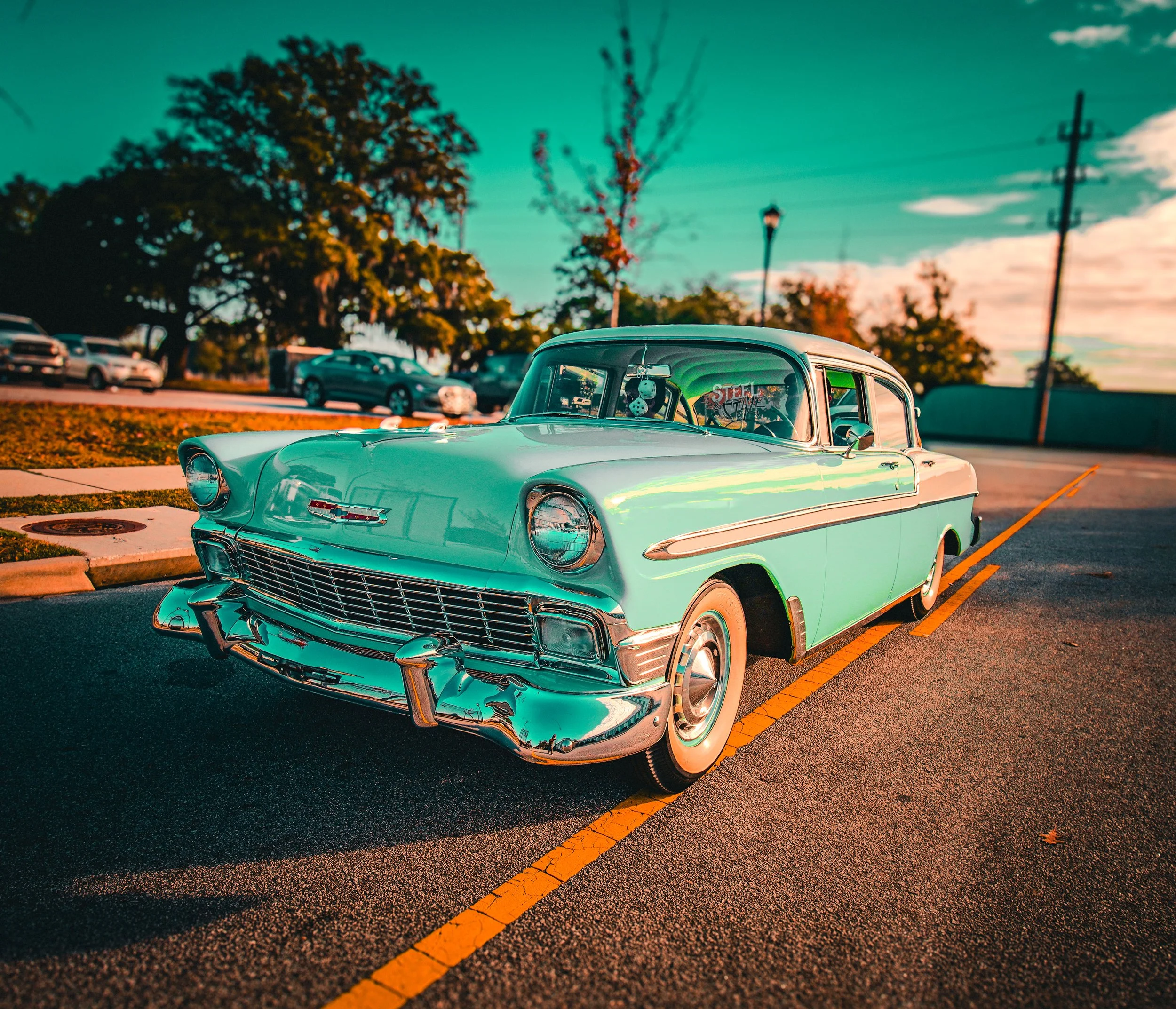 A vintage light blue Chevrolet car parked in a parking lot during sunset with other cars and trees in the background.