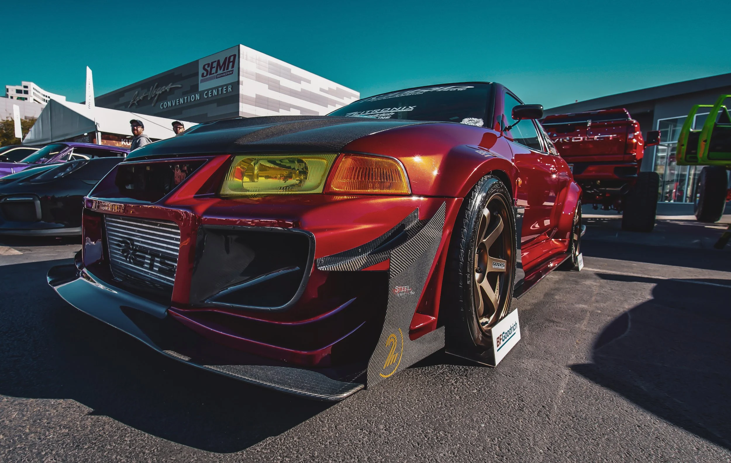 A red race car with custom modifications, including a carbon fiber front splitter, racing wheels, and greenish-yellow headlights, displayed at an auto show with other cars and a convention center in the background.