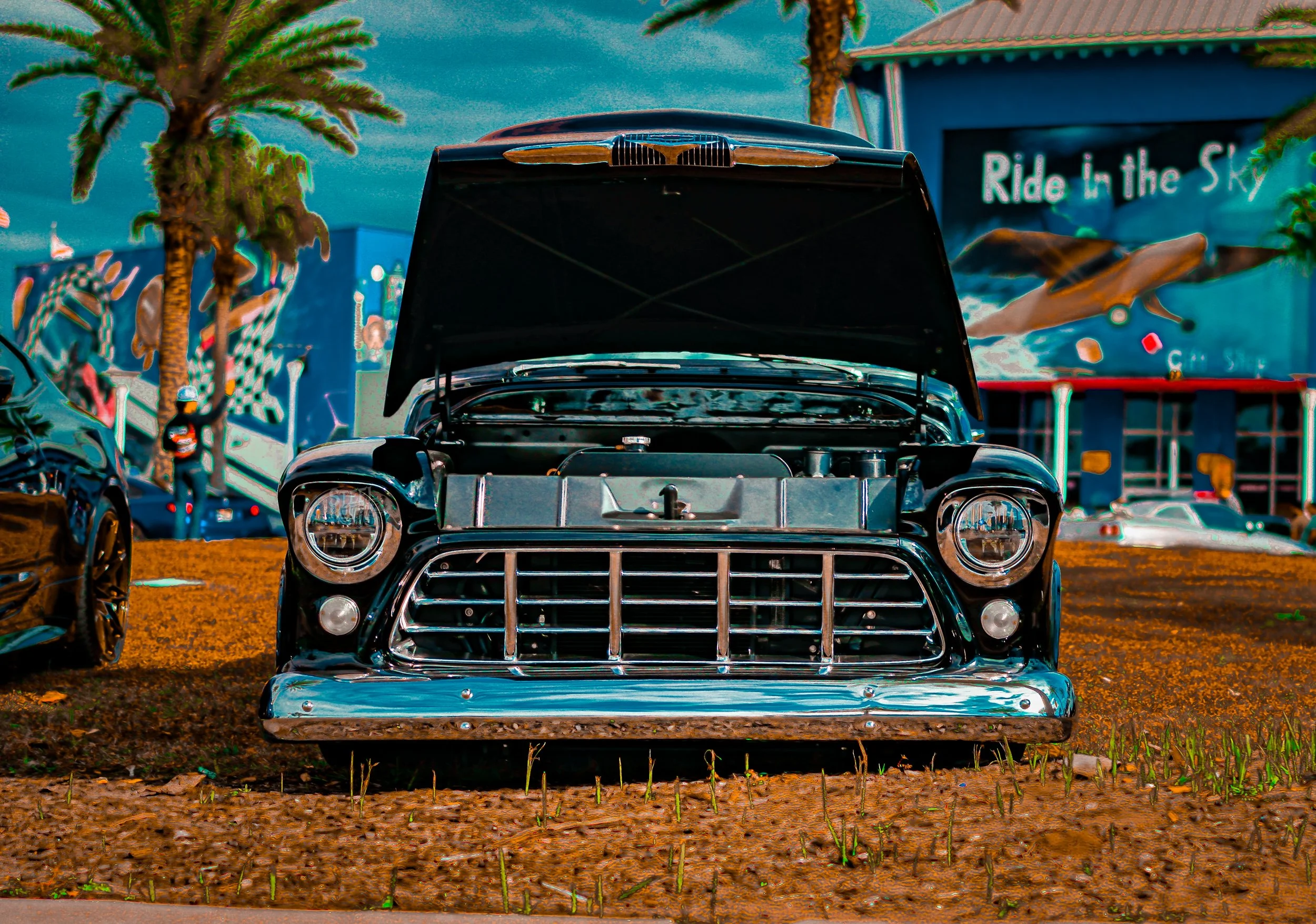 A classic black car with its hood open, parked on dirt, with palm trees and a mural with a whale and the phrase "Ride in the Sky" in the background.