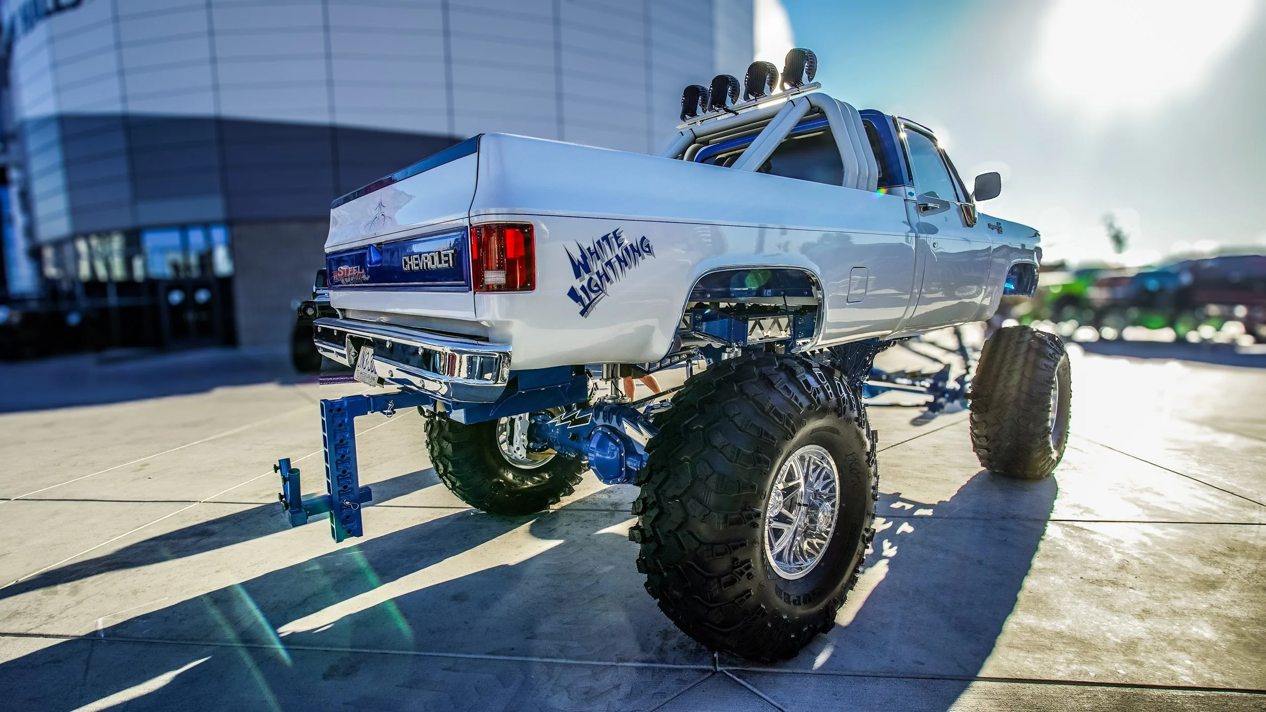 A custom white monster truck with large off-road tires and lifted suspension, parked outdoors in front of a modern building, with the words "White Lightning" written on the side.