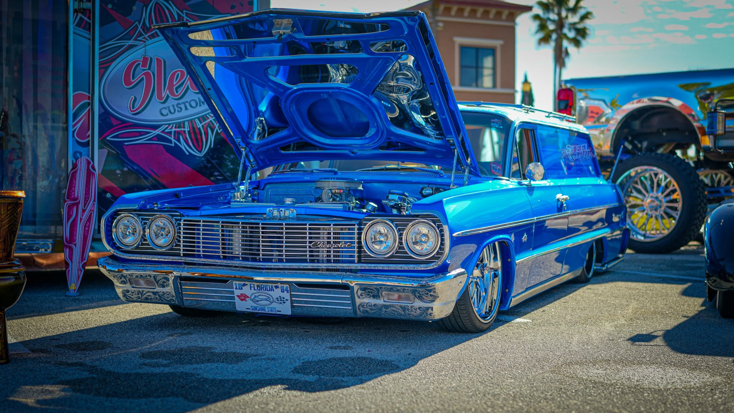 A vintage blue Chevrolet lowrider with a lowered stance and chrome accents is on display at a car show, with its hood open revealing the engine. The car has custom wheels and an Florida license plate that reads 'Sunshine State'.