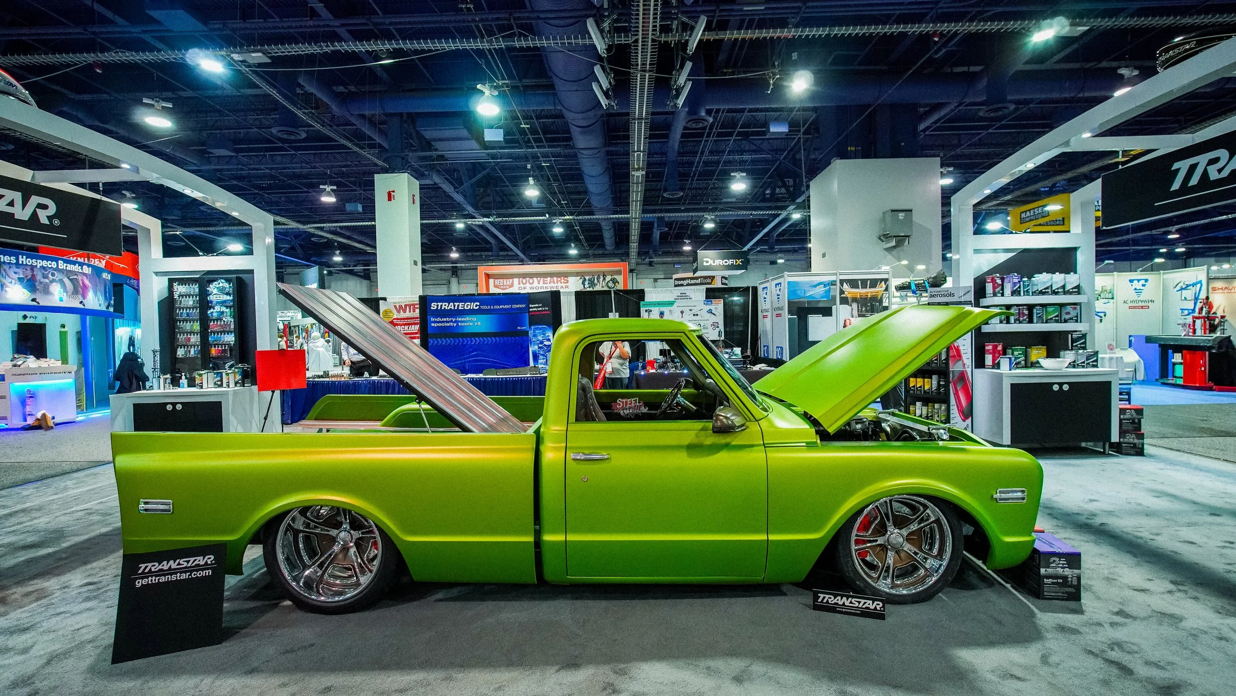 Bright green vintage pickup truck with lowered stance and large custom chrome wheels at an indoor auto show, with its hood and tailgate open.