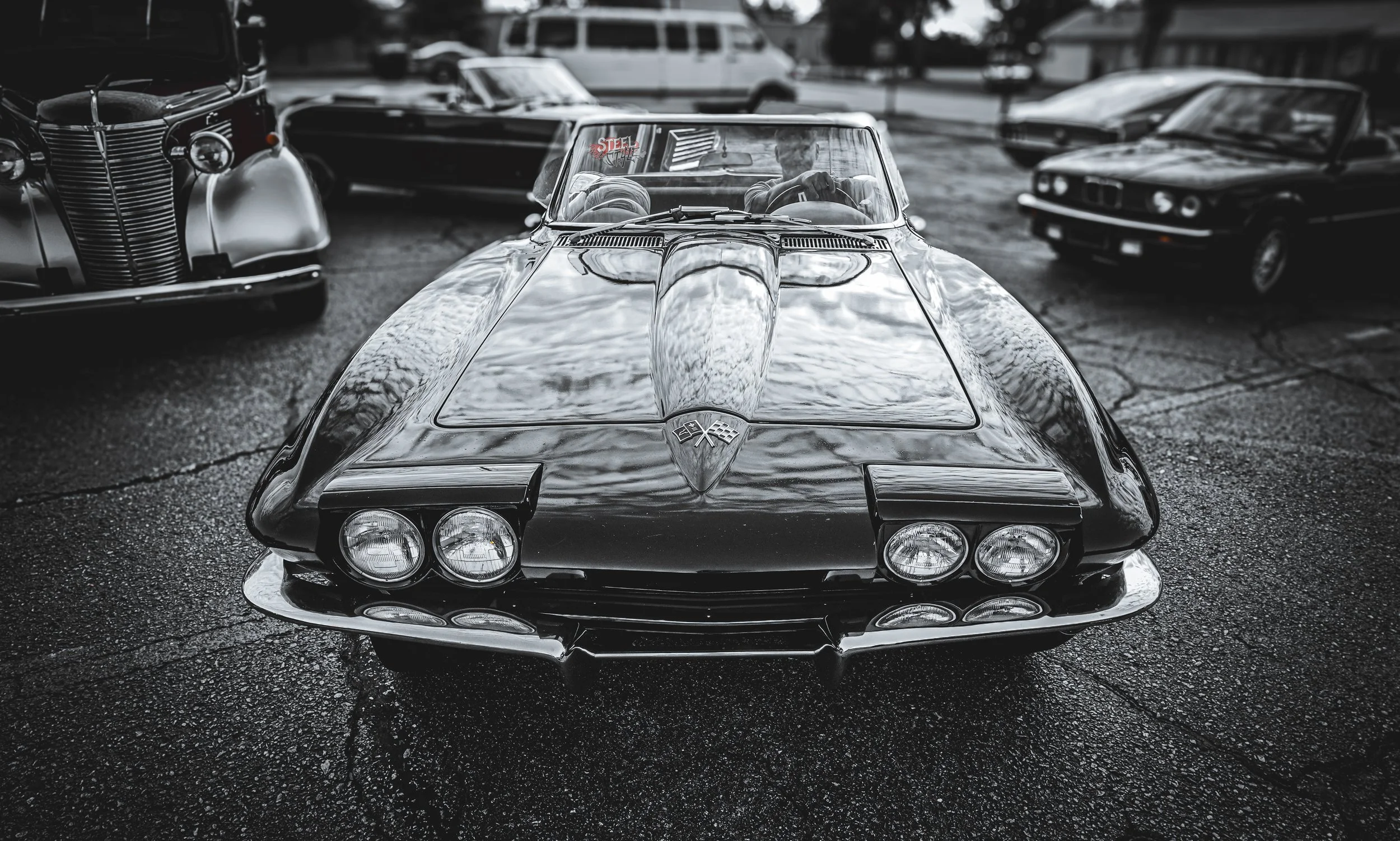 A black vintage Chevrolet Corvette car with a sleek, reflective surface, parked in an outdoor lot among other classic cars, with a man inside