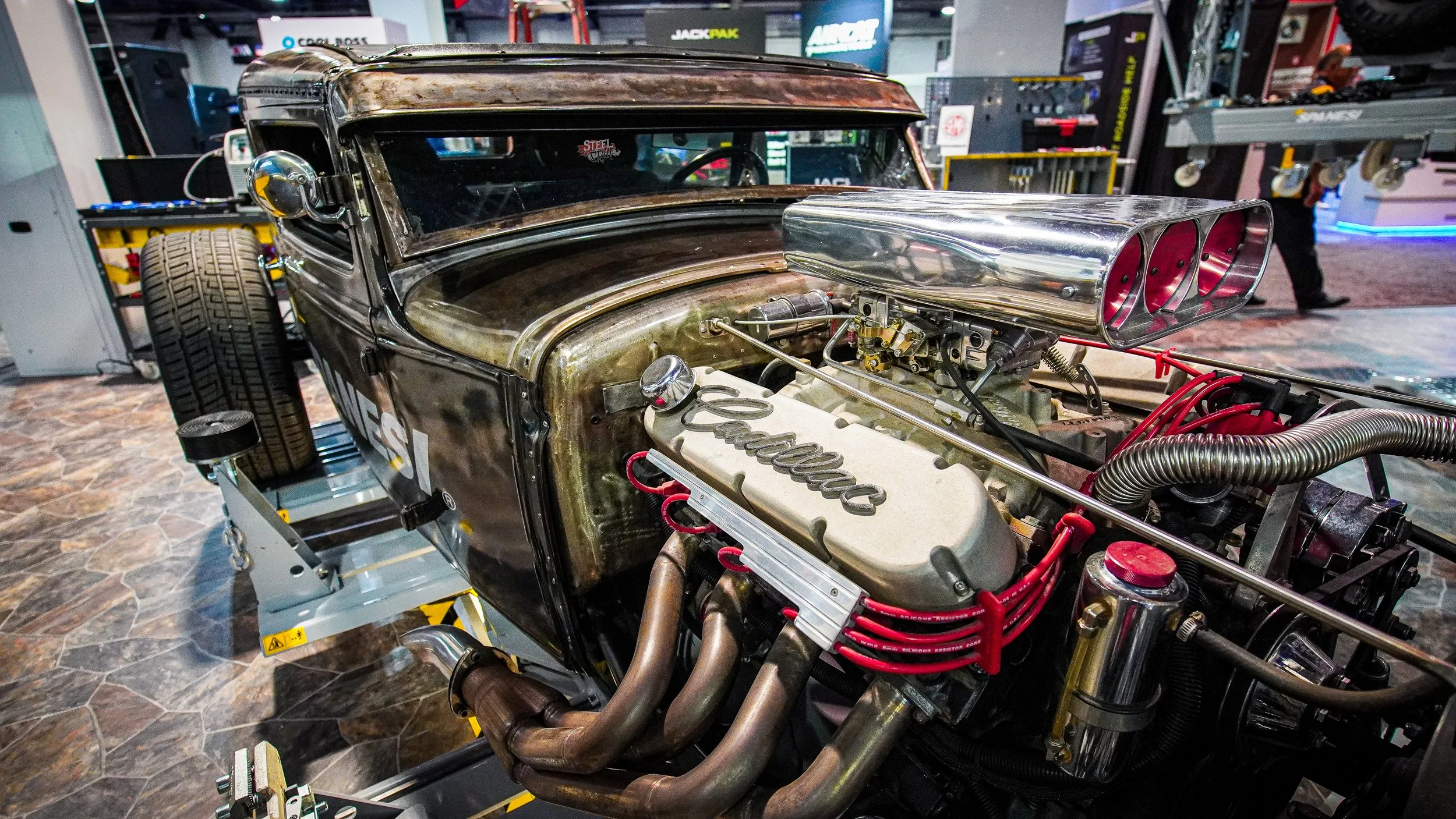 A vintage car with an exposed engine on display, featuring a supercharger with three intake openings, red spark plug wires, and a brass engine cover with the word 'Edelbrock' on it. The body of the car is black with a weathered finish, and it is posi