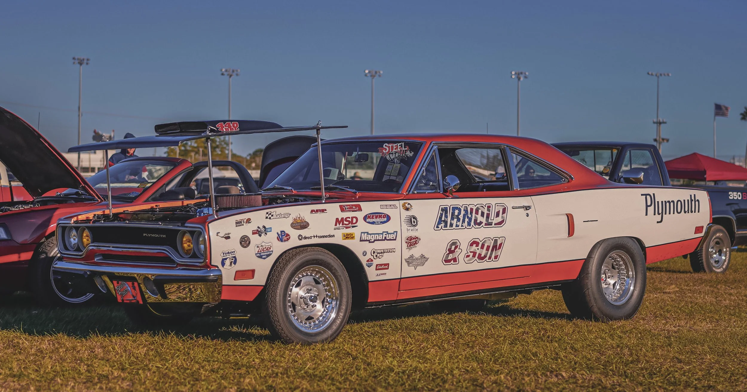 A vintage Plymouth racing car with a white and red paint job, featuring various sponsor logos, parked on grass at a racing event, with other cars, tent, and flags in the background.
