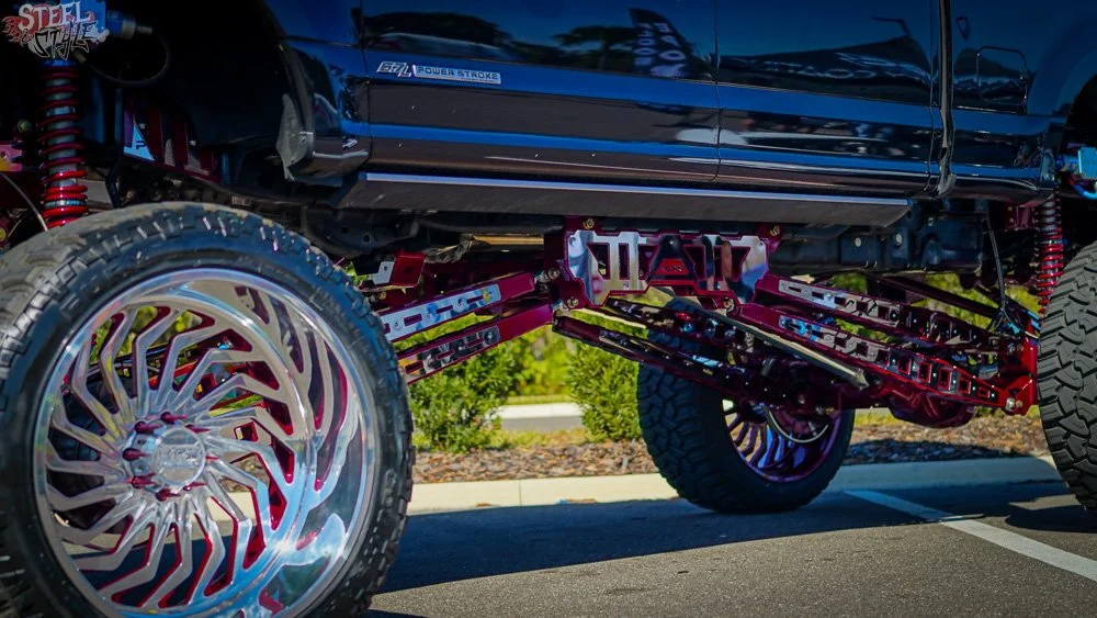Close-up of customized off-road truck with large chrome wheels, suspension system, and red undercarriage parts parked in a lot.