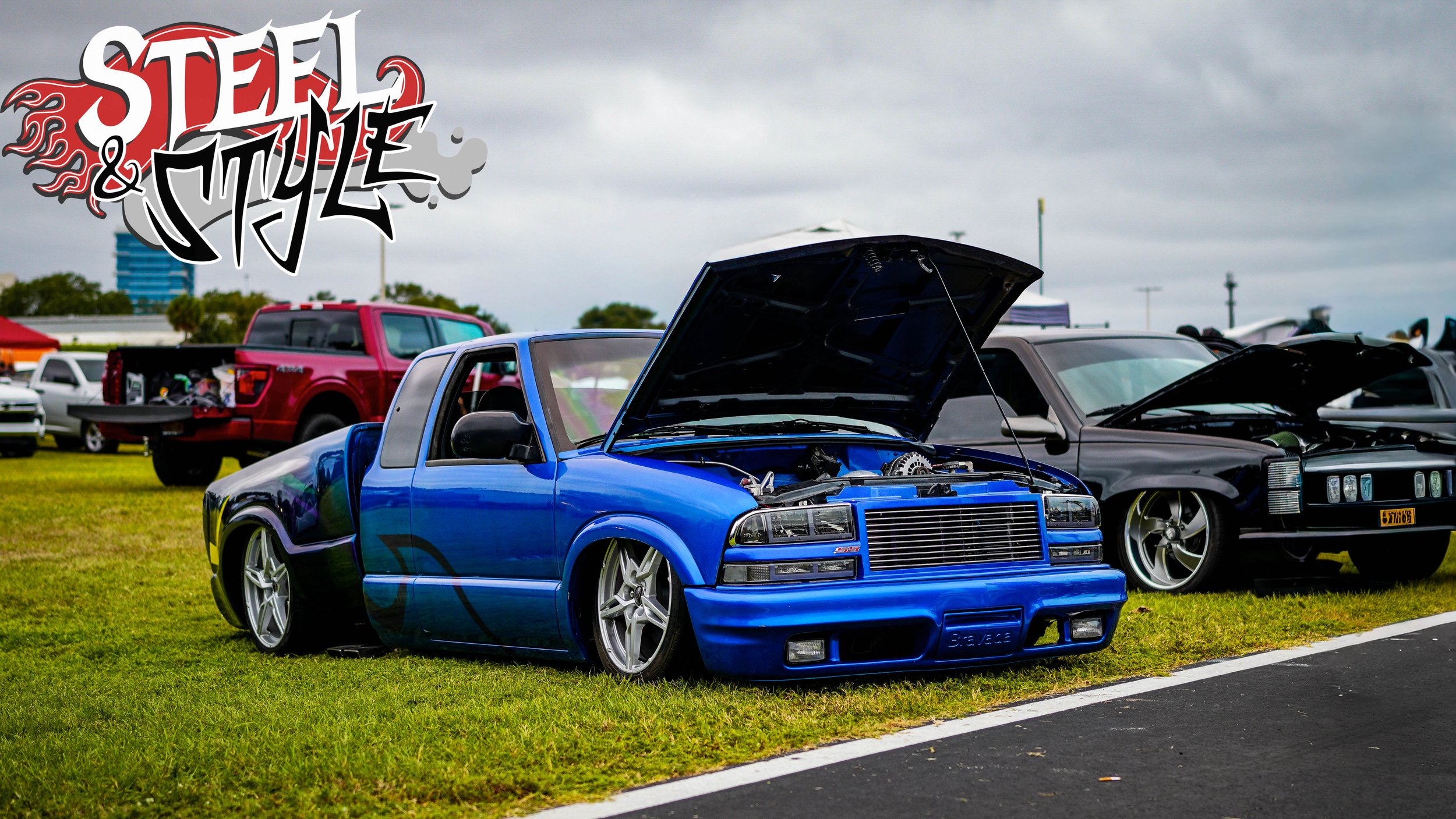 A modified blue pickup truck with its hood open, parked on a grass field at a car show, with other cars in the background and a logo that reads "Steel & Style" in the upper left corner.