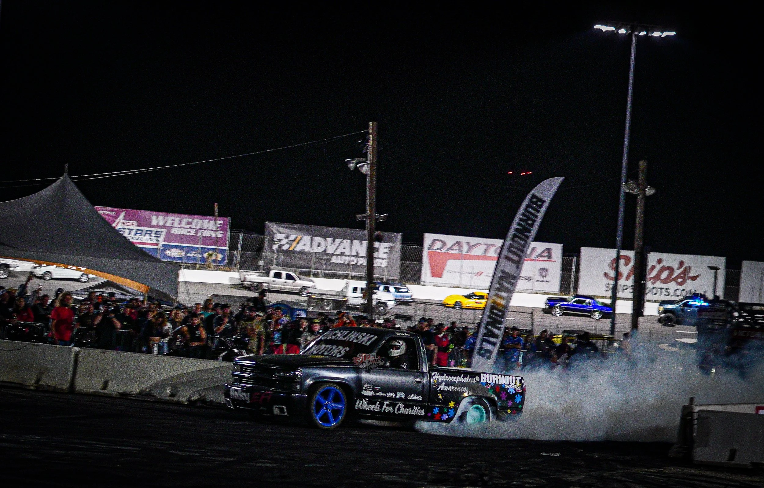 Nighttime race track scene with a black pickup truck doing burnout, surrounded by smoke and a crowd of spectators behind barriers, with car advertisements and banners in the background.