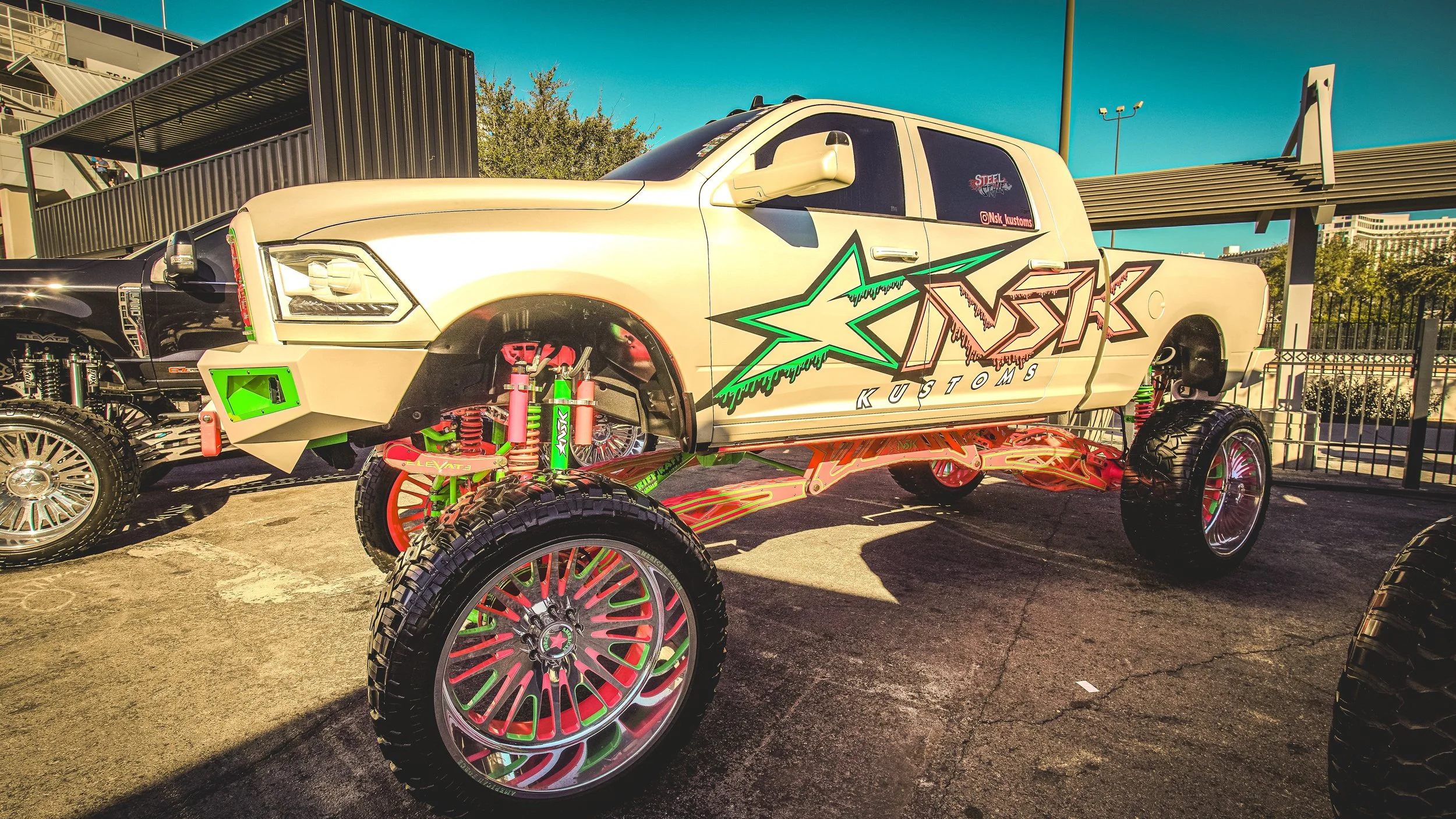 A heavily modified pickup truck with a large, custom body featuring a 'STOCK KUSTOMS' logo, lifted high on altered suspension with large, colorful wheels and off-road tires, parked in a lot with other vehicles and structures in the background.