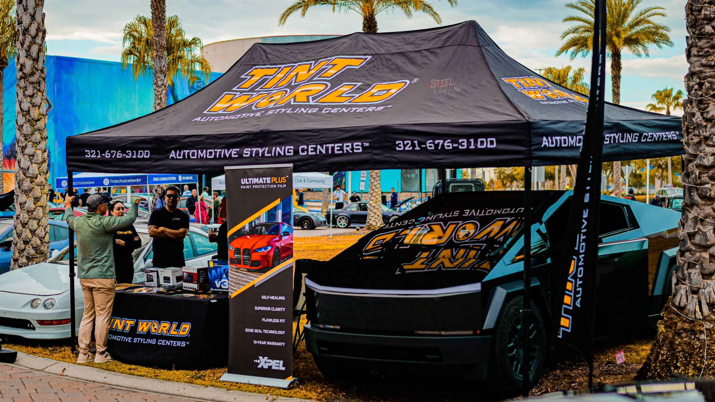 A tent at an outdoor event with the logo of 'Tint World Automotive Styling Centers,' a car is parked inside the tent along with a display table, and palm trees line the background.