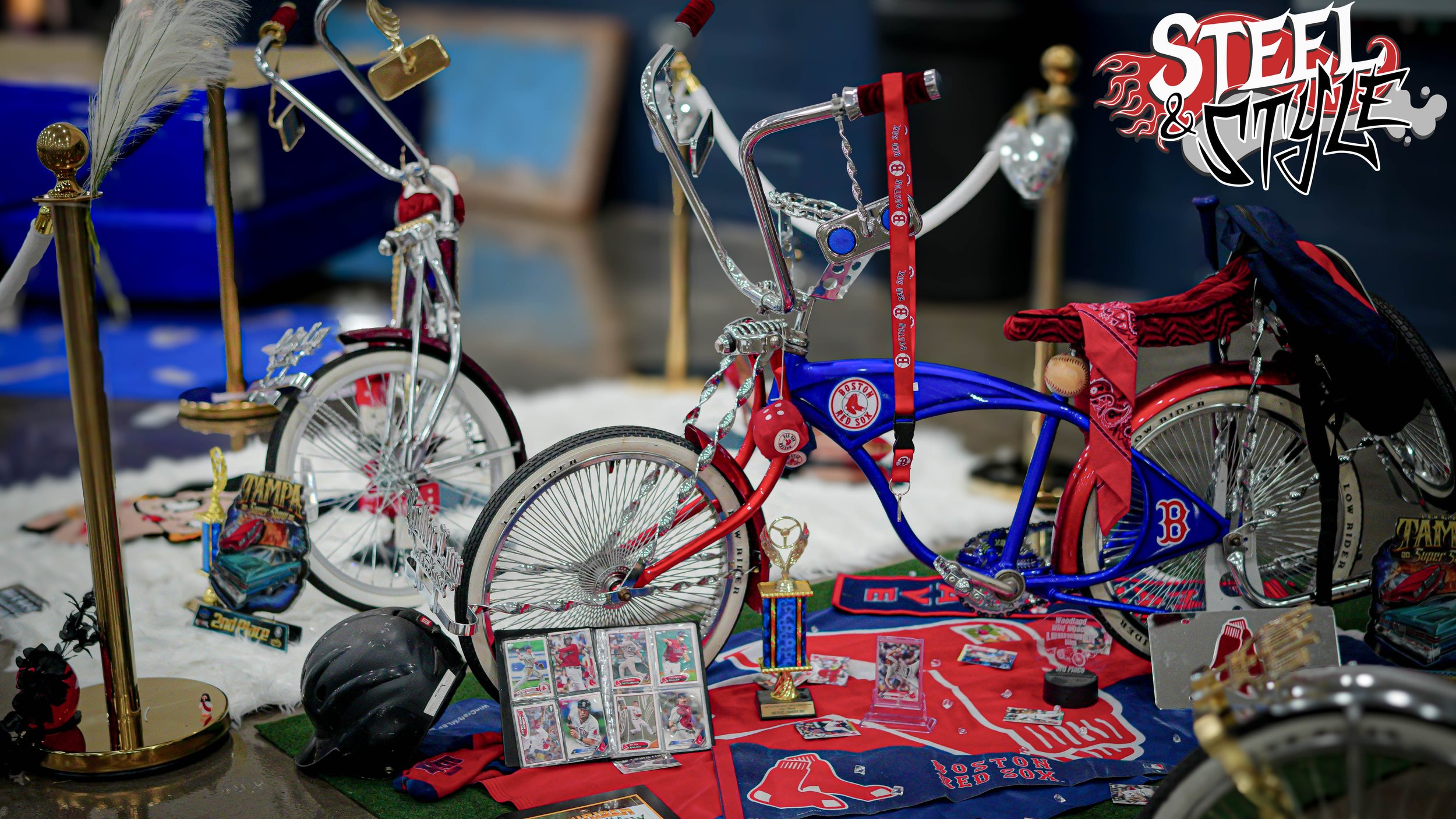 A display of Boston Red Sox themed bicycle and memorabilia, including a blue and red bike, sports cards, trophies, and team gear, set on a snow-like surface.