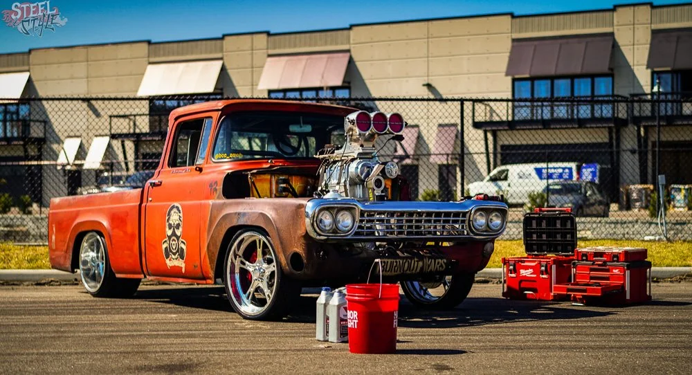 A modified vintage pickup truck with a large supercharged engine and racing decals, parked on the street with tools and supplies nearby.