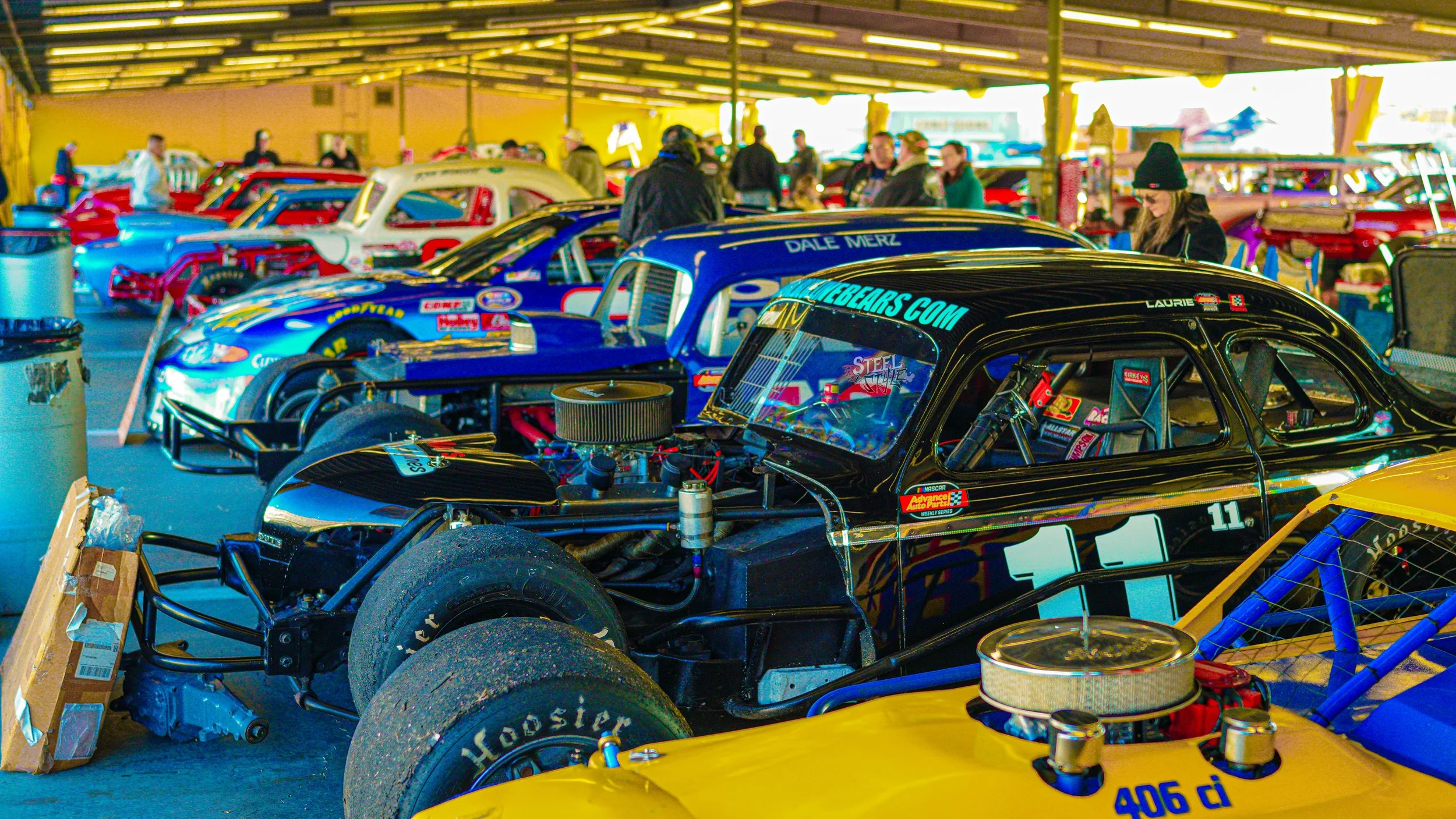 Several race cars parked inside a covered garage area, with people walking and inspecting the vehicles.