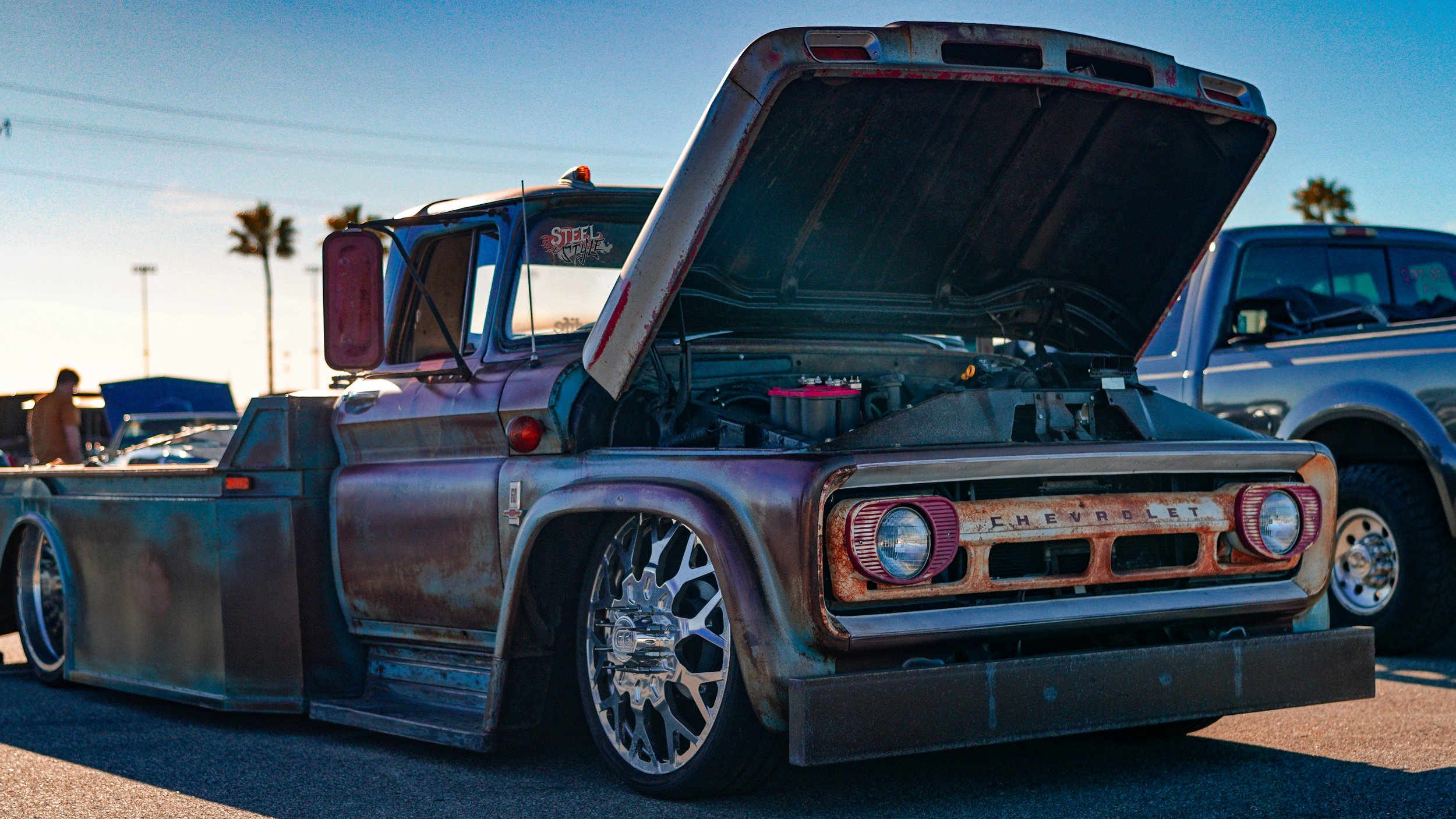 Rusty Chevrolet pickup truck with lowered stance and large custom wheels, hood open showing engine, at outdoor car show.