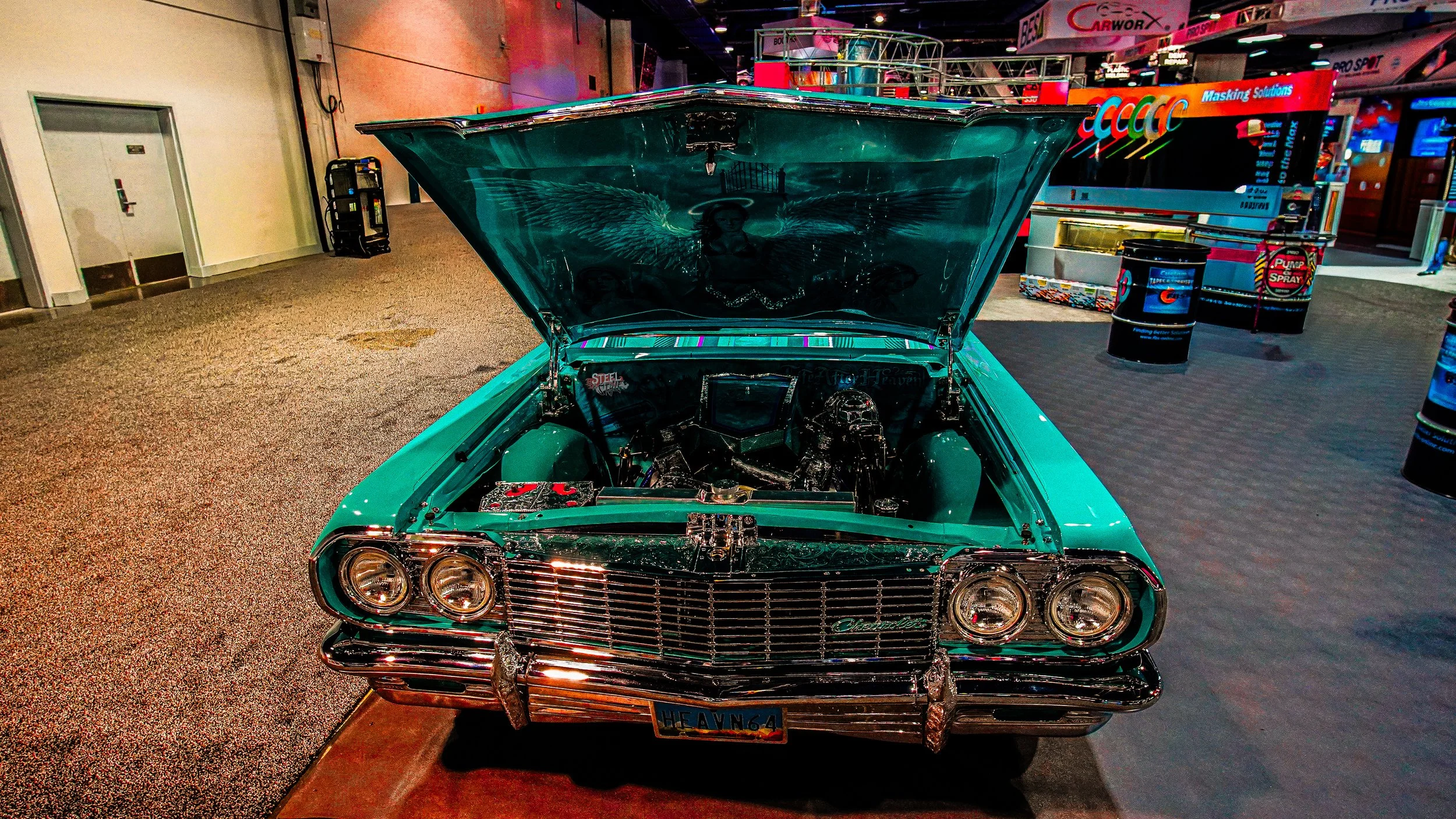 Front view of a vintage teal-colored Chevrolet car with its hood open, showcasing the engine, displayed indoors at an event or exhibition.