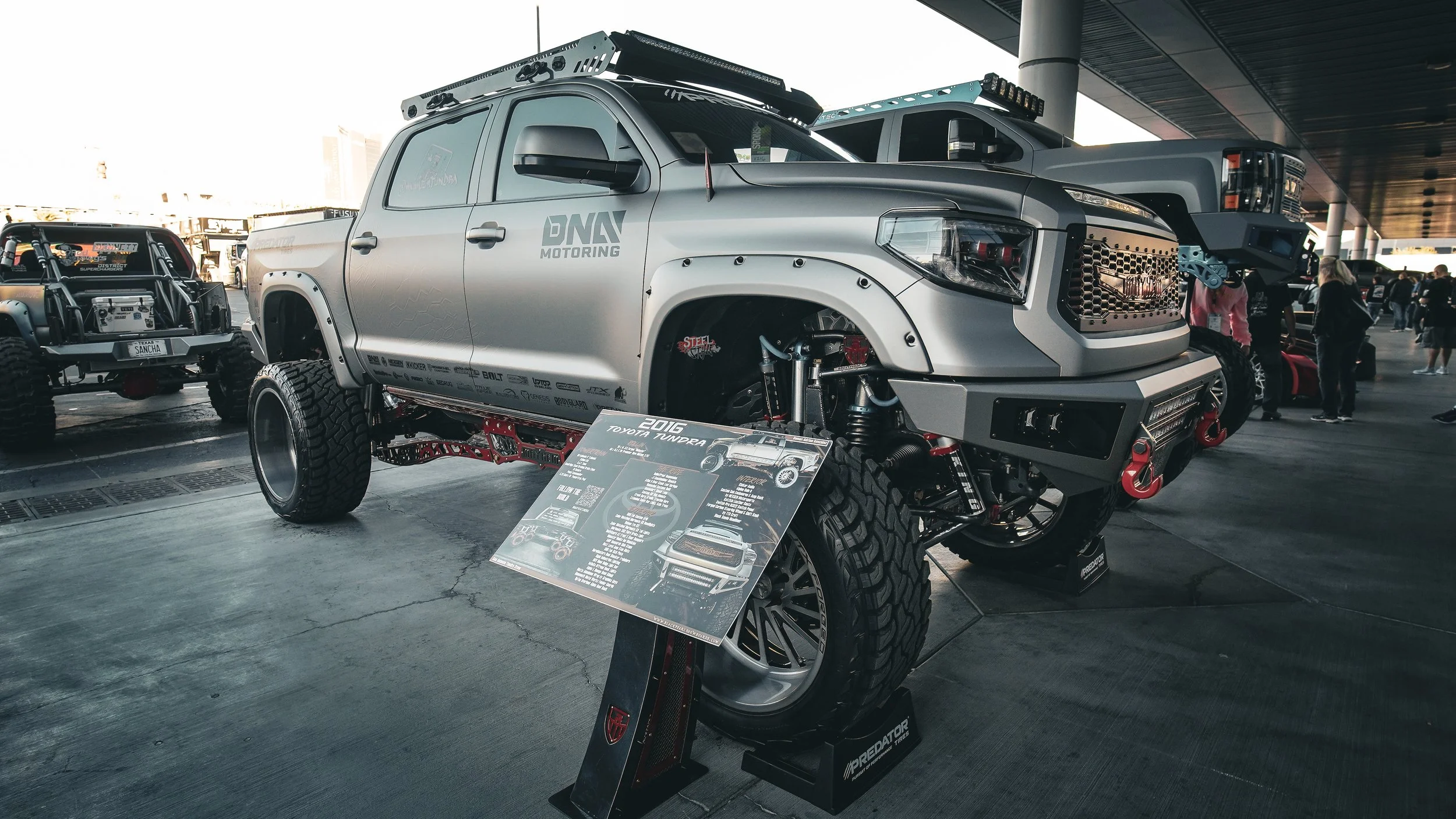 A modified silver Toyota Tundra pickup truck with large off-road tires and suspension, displayed at an auto show with a signboard in front providing specifications and details.