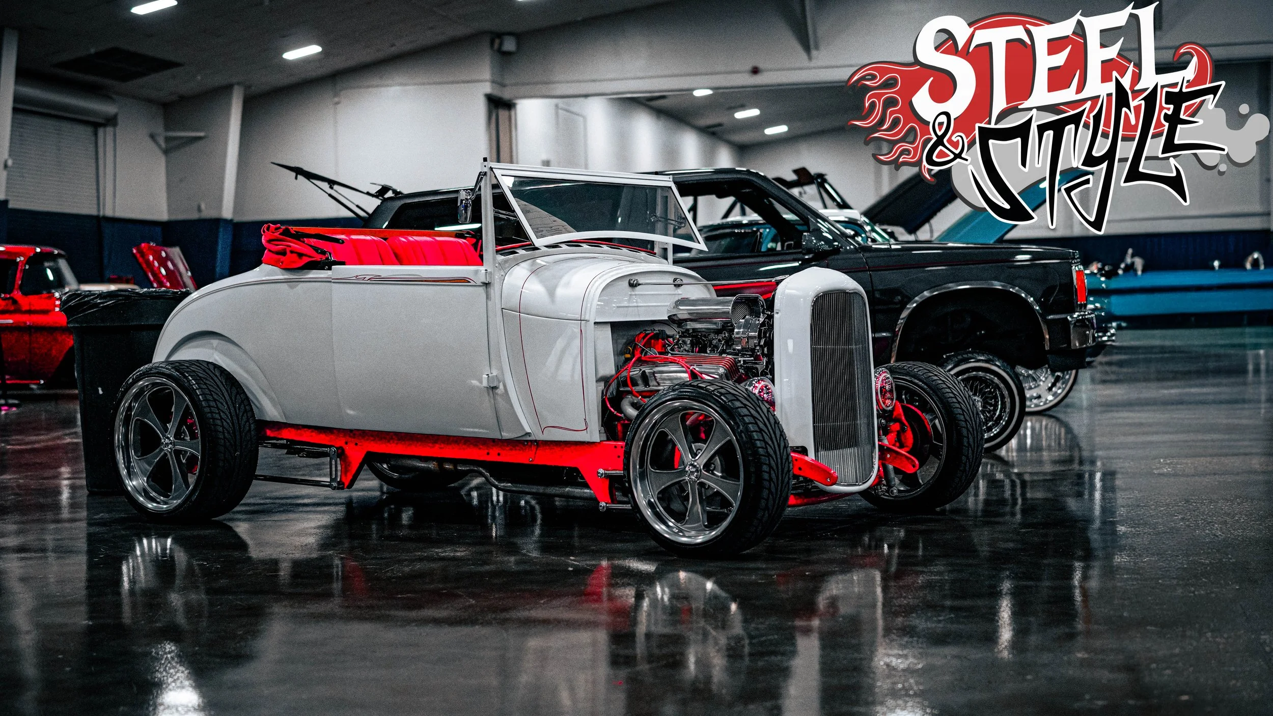 A vintage car with a white body and red interior on display at an indoor car show, with other classic cars in the background and a "Steel & Style" logo in the top right corner.