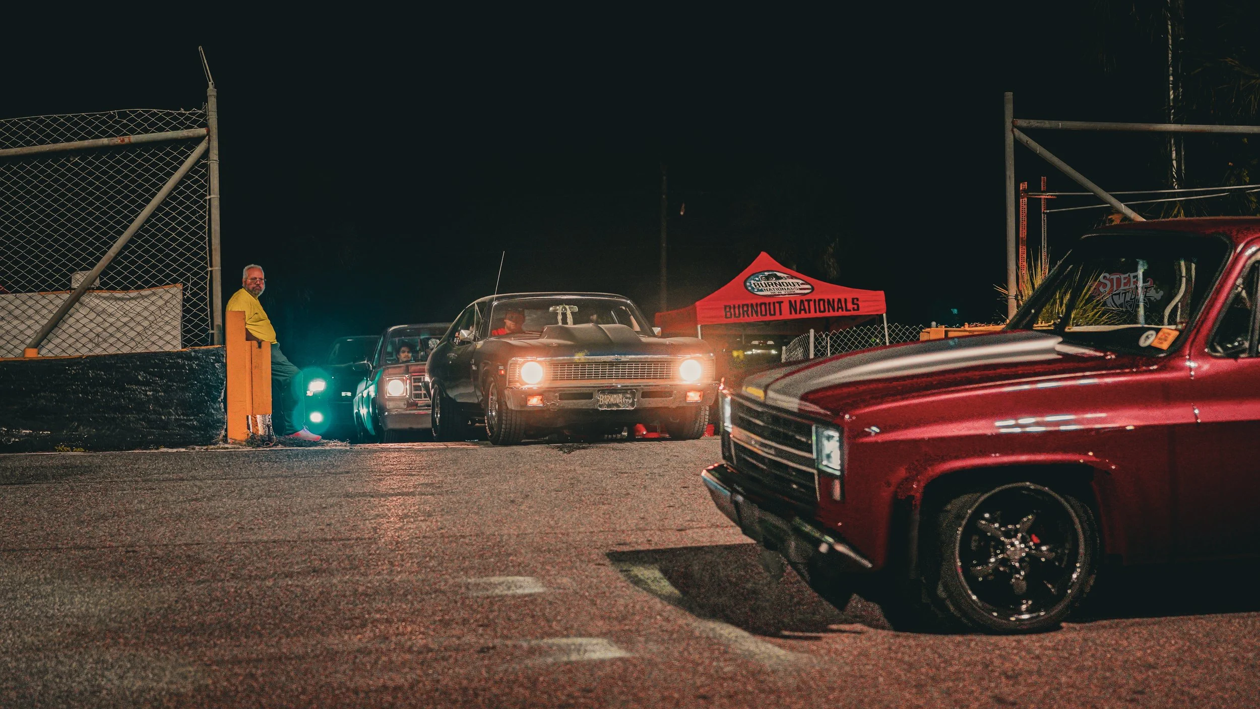 Nighttime scene of vintage cars parked near a chain-link fence with a man in a yellow jacket leaning against a pole, and a red tent labeled 'BURNOUT NATIONALS' in the background.