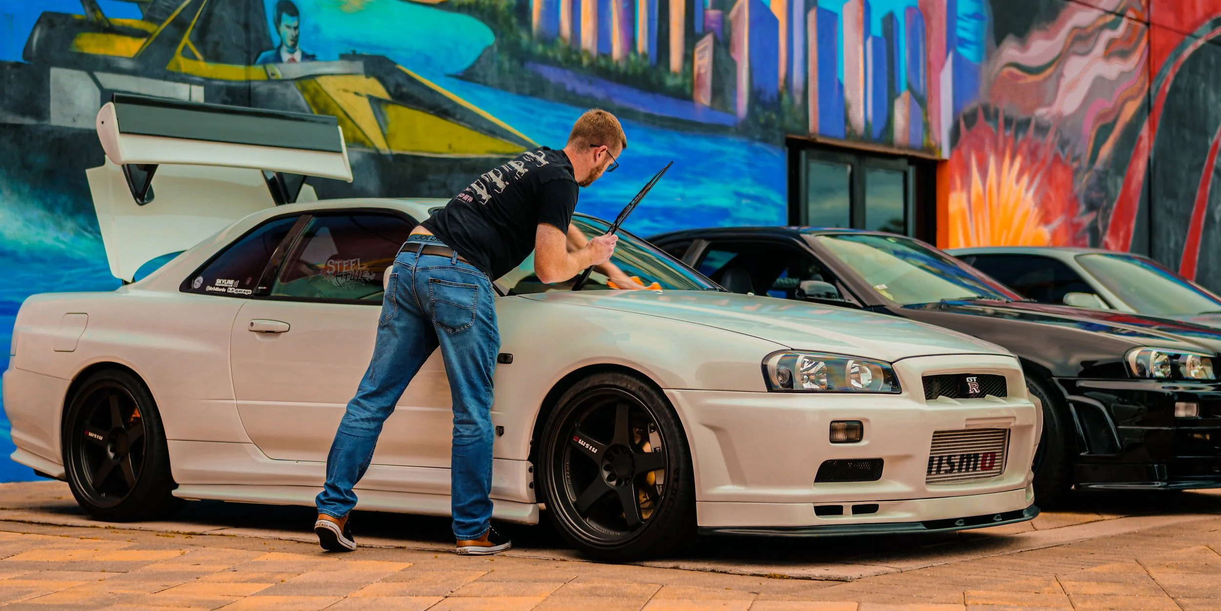 A man in jeans and a black T-shirt cleaning a white Nissan Skyline GT-R R32 with a cloth, parked inside a garage with vibrant street art murals, alongside other sports cars.