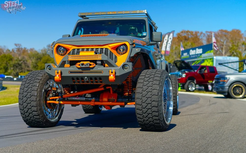 A highly modified off-road vehicle with large tires, a custom suspension, and a rugged front bumper, parked on a racing track with other vehicles and banners in the background.