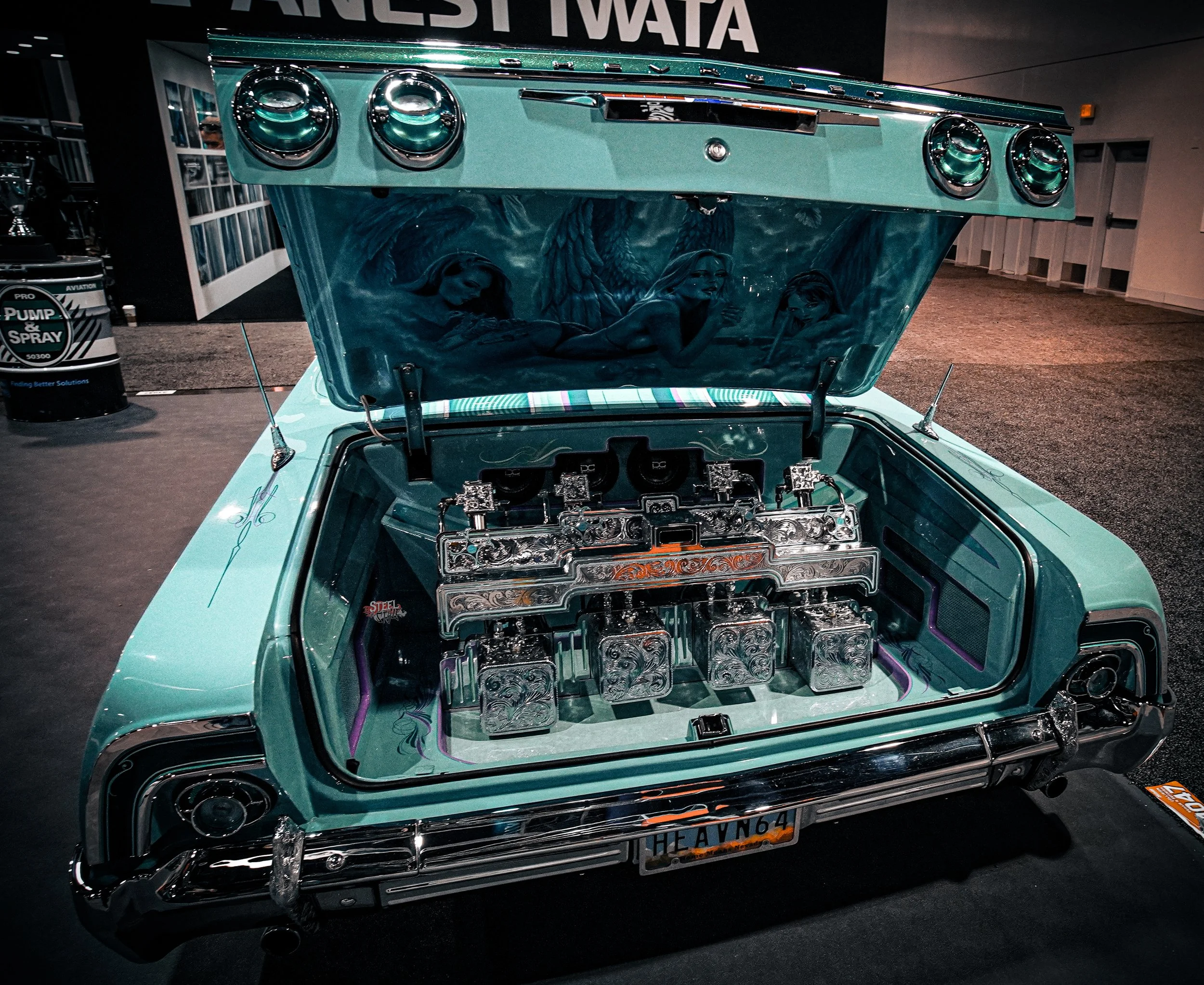 The front of a vintage car with its hood open, displaying a highly detailed and ornate engine with engravings. The inside of the hood features painted artwork of three women with angel wings.