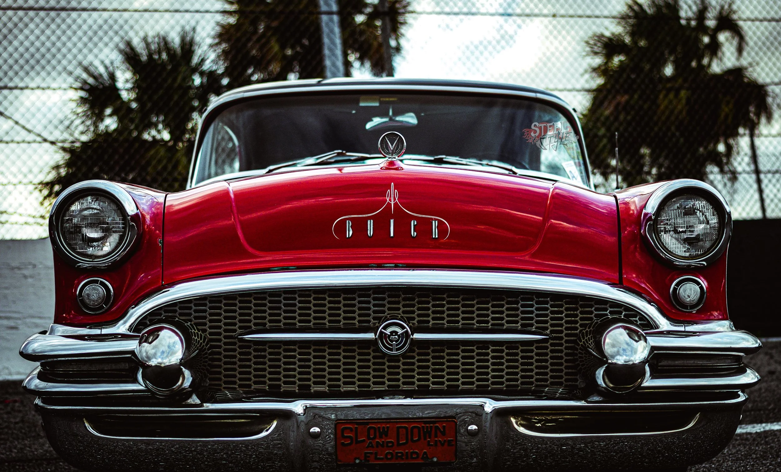 Front view of a vintage red Buick car with a chrome grille and round headlights, parked behind a chain-link fence.