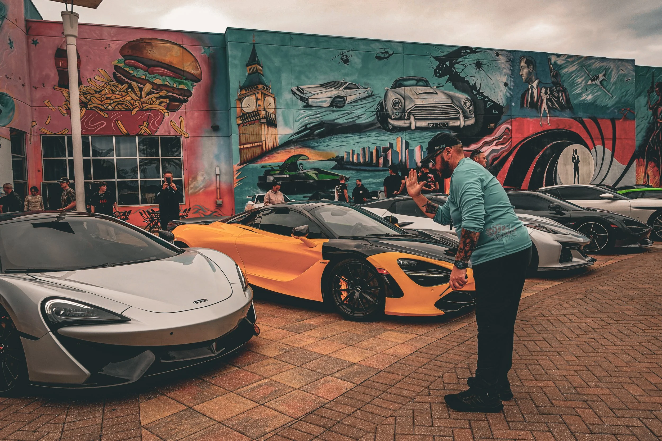 A man with tattoos and a cap is giving a high five to a person outside a parking lot filled with luxury sports cars, including McLarens and Lamborghinis, in front of a colorful mural depicting various images like a hamburger, Big Ben, classic cars, h
