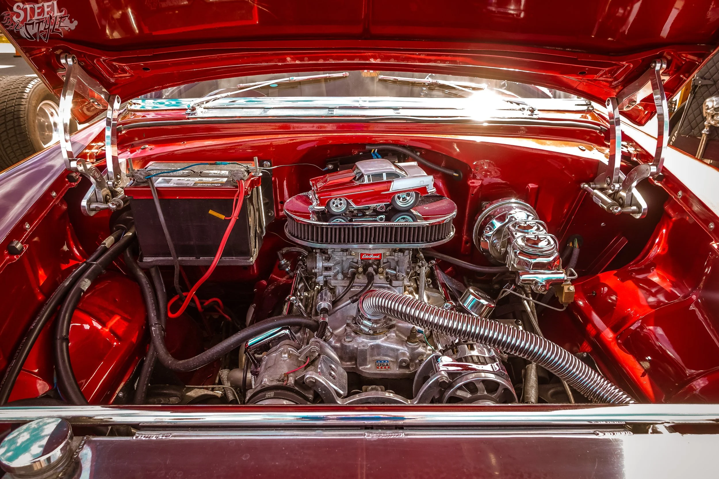 View of a classic red car engine bay with chrome and black components, including a small toy car on top of the engine.