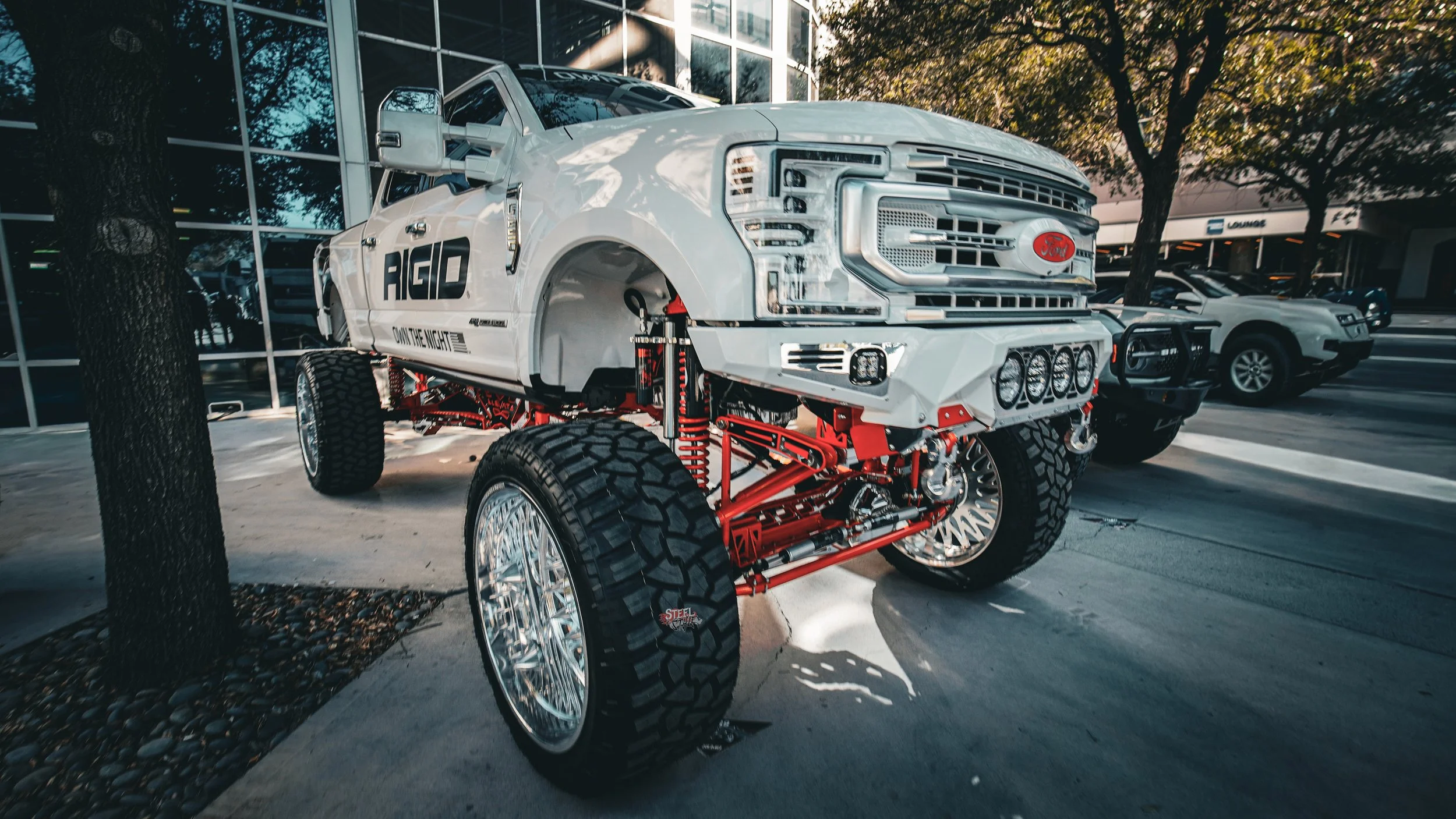 A modified Ford truck with oversized chrome wheels, large off-road tires, and a lifted suspension on display outdoors near trees and a glass building.