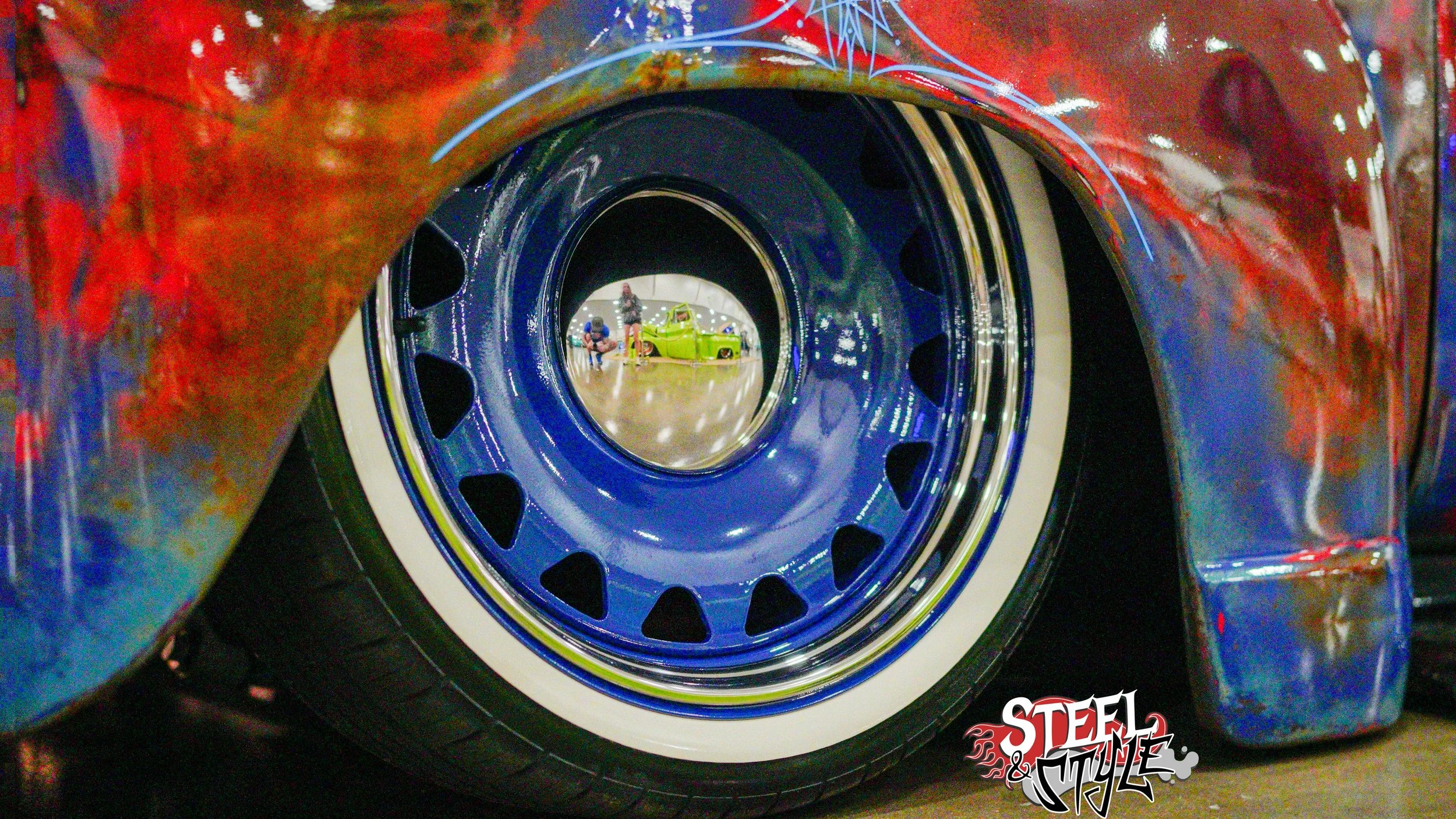Close-up view of a car wheel with colorful body paint and a reflection of people and a vintage green car in the shiny hubcap.