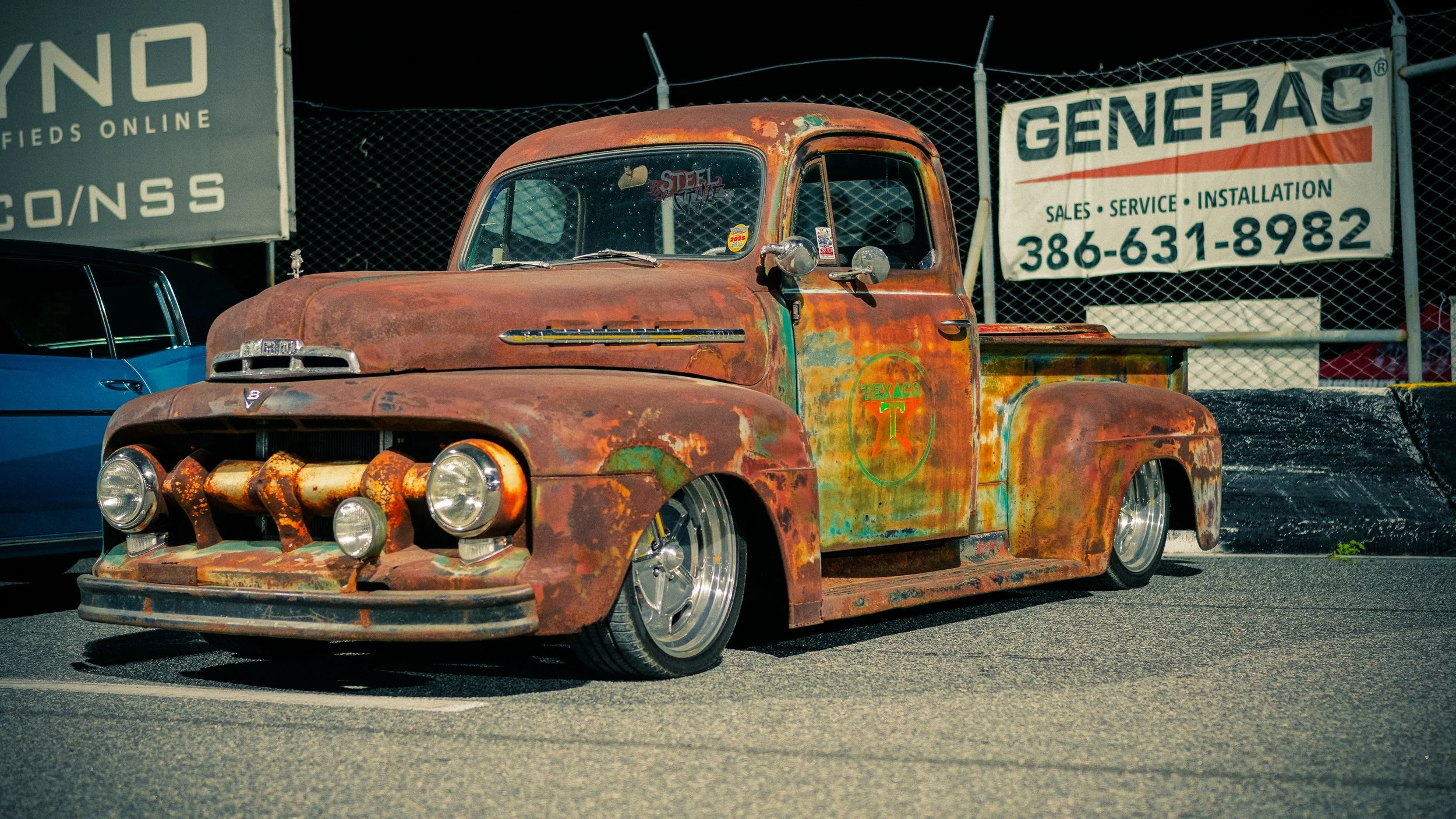 Rusty vintage pickup truck with lowered wheels parked on street at night, with signs for Generac and Texaco in the background.