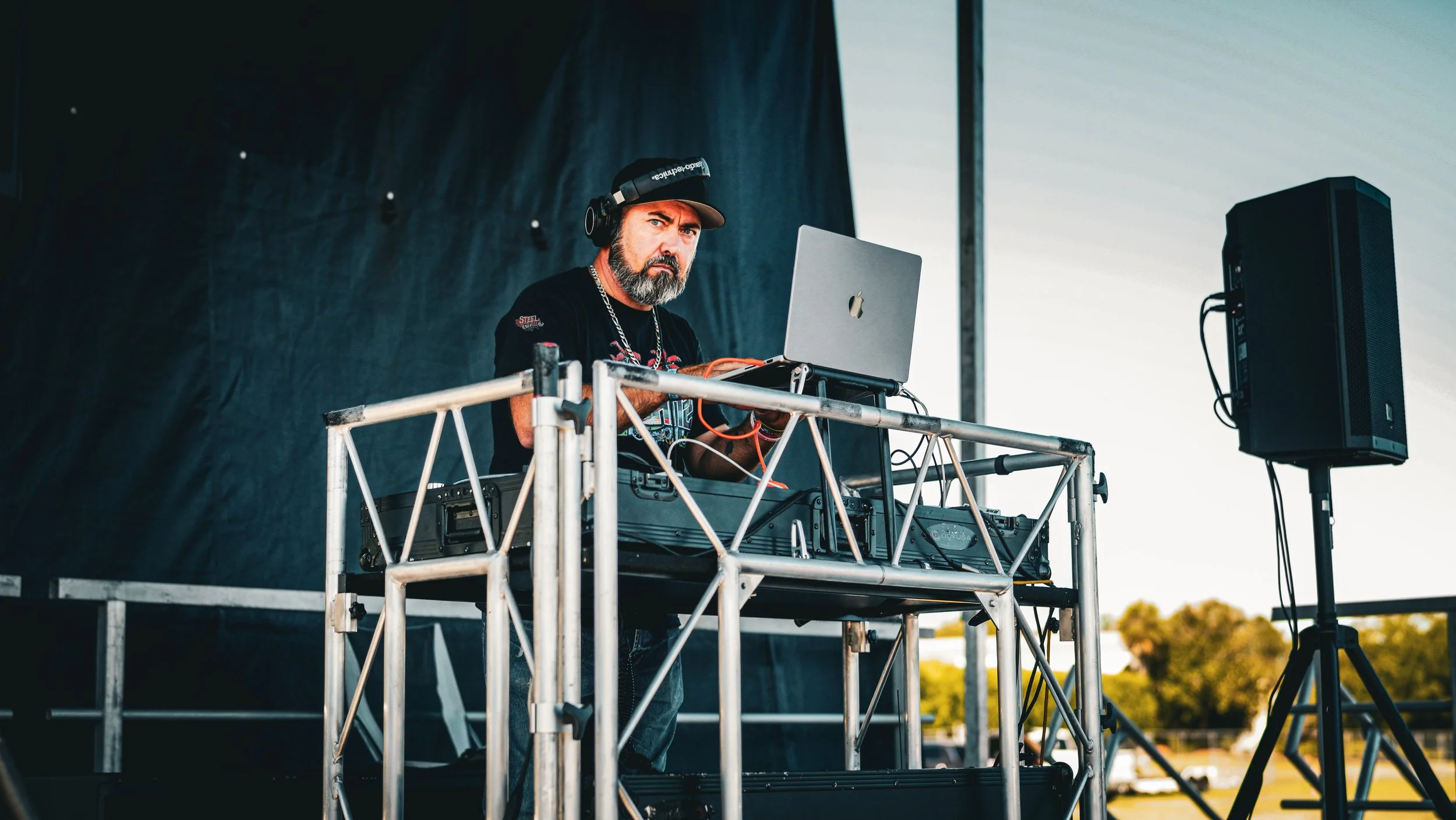 A DJ with a beard and headphones, wearing a black t-shirt with a chain necklace, performing at an outdoor event. He is standing behind a DJ setup on a metal platform, with a laptop and sound equipment. A large speaker is positioned to his right, and 