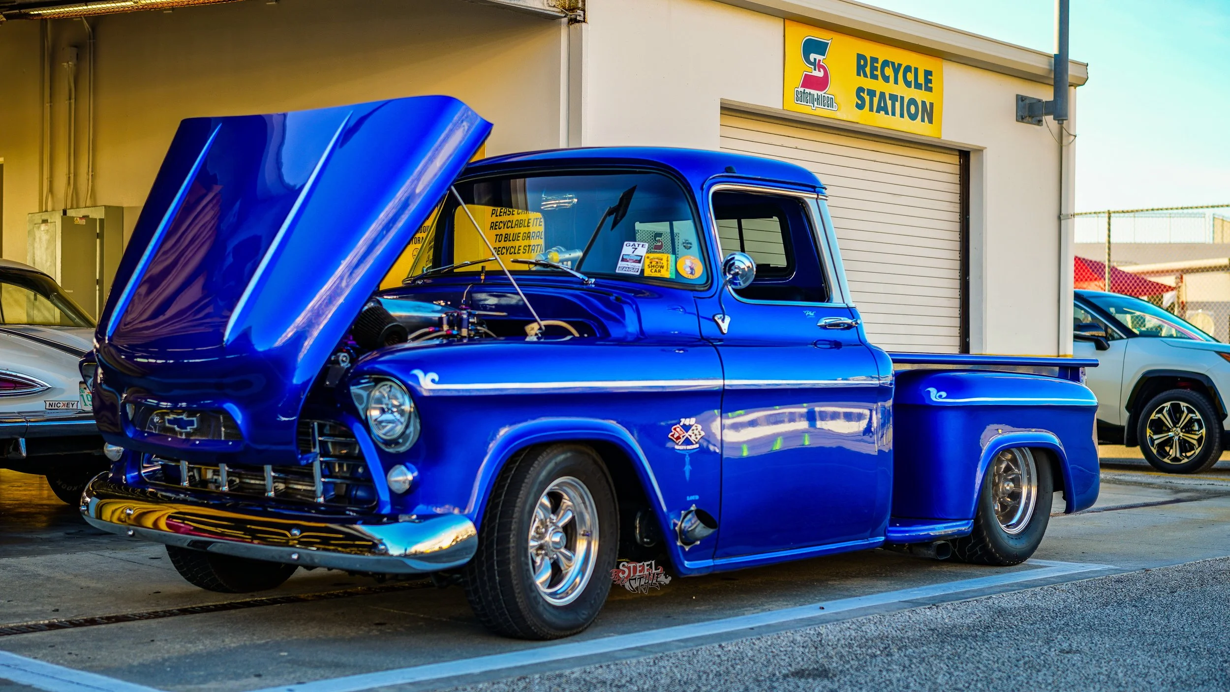A vintage blue pickup truck with its hood open parked outside a recycle station. The truck has a shiny chrome bumper and wheel rims, with a small logo and decals on the side. Other cars are visible nearby.
