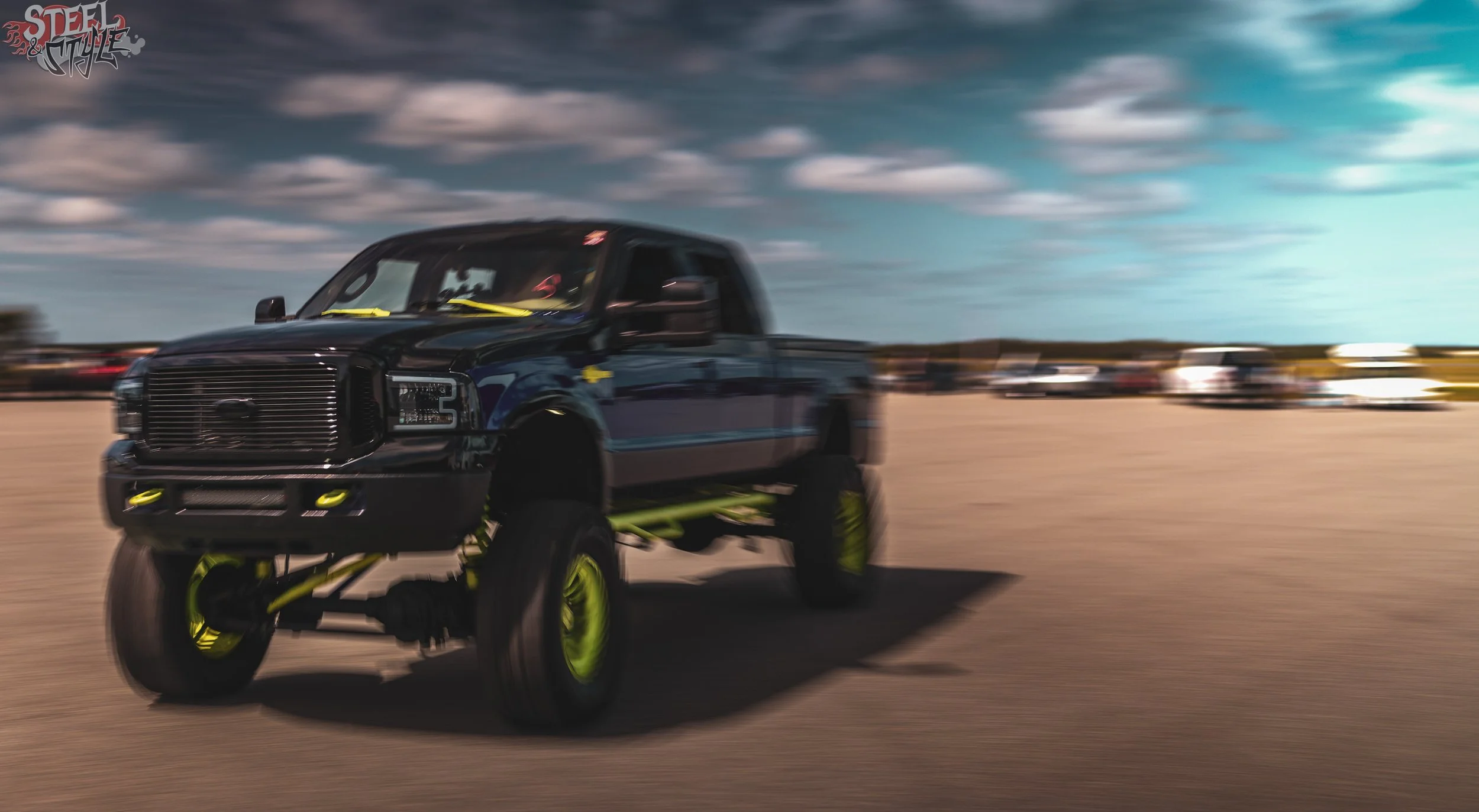 A black monster truck with neon yellow wheels driving across a desert with blurred cars and a cloudy sky in the background.