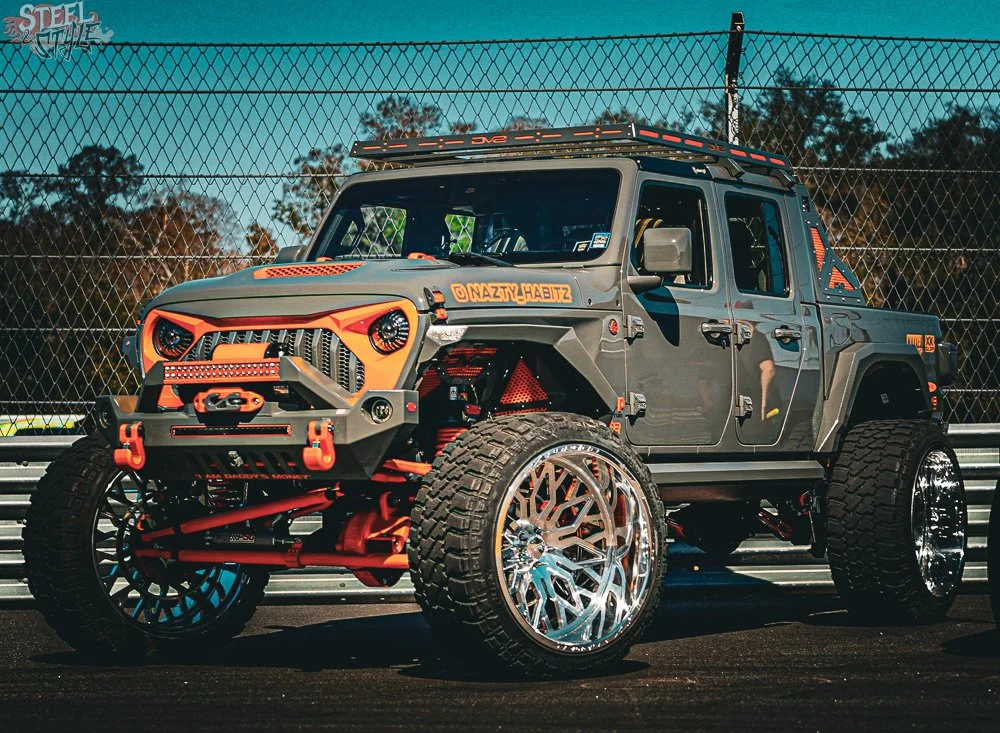 A custom modified off-road pickup truck with large wheels, orange accents, and a rugged design, parked on a race track against a chain-link fence.